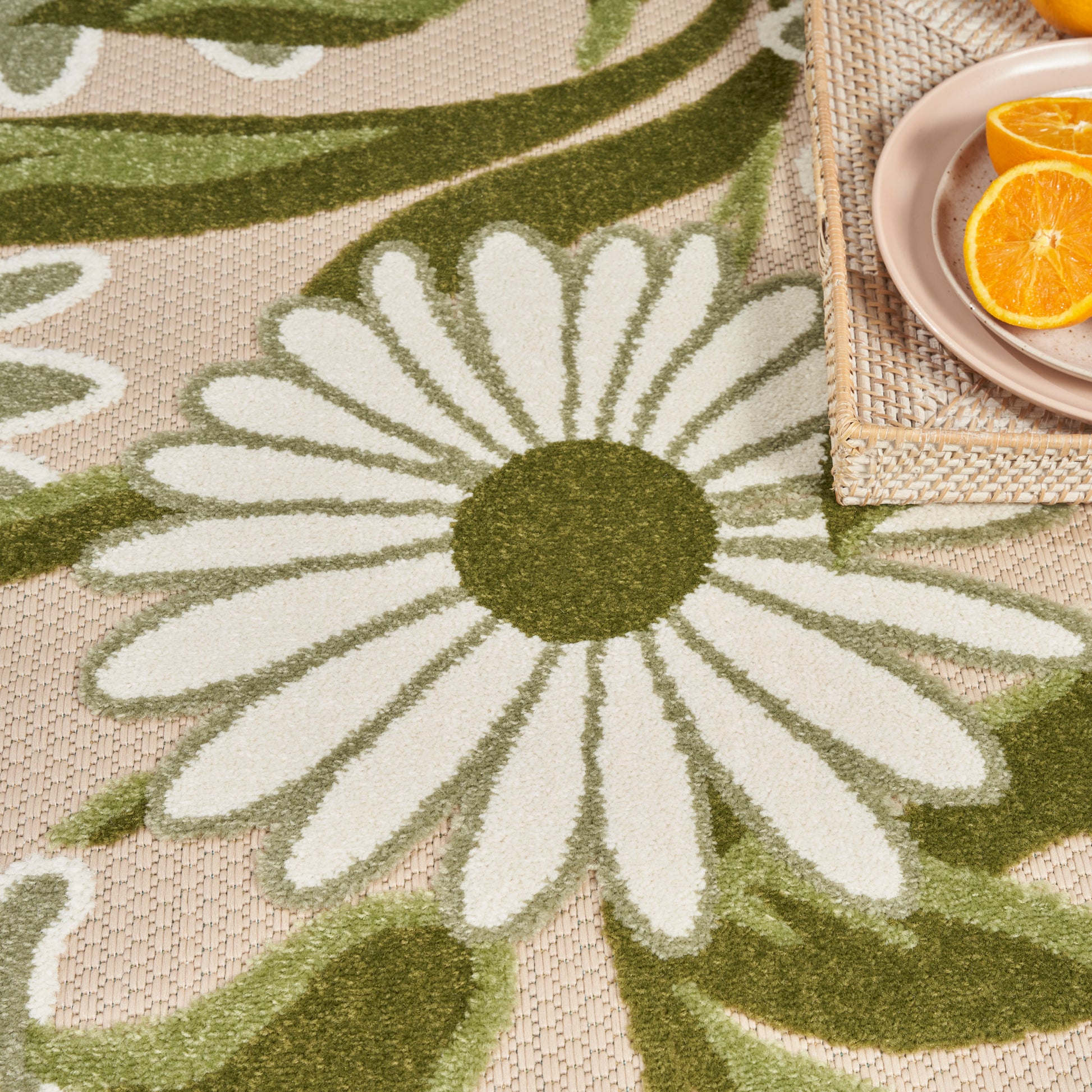 Beige woven rug featuring a white daisy with green center; natural woven tray holding a pink plate with orange slices.