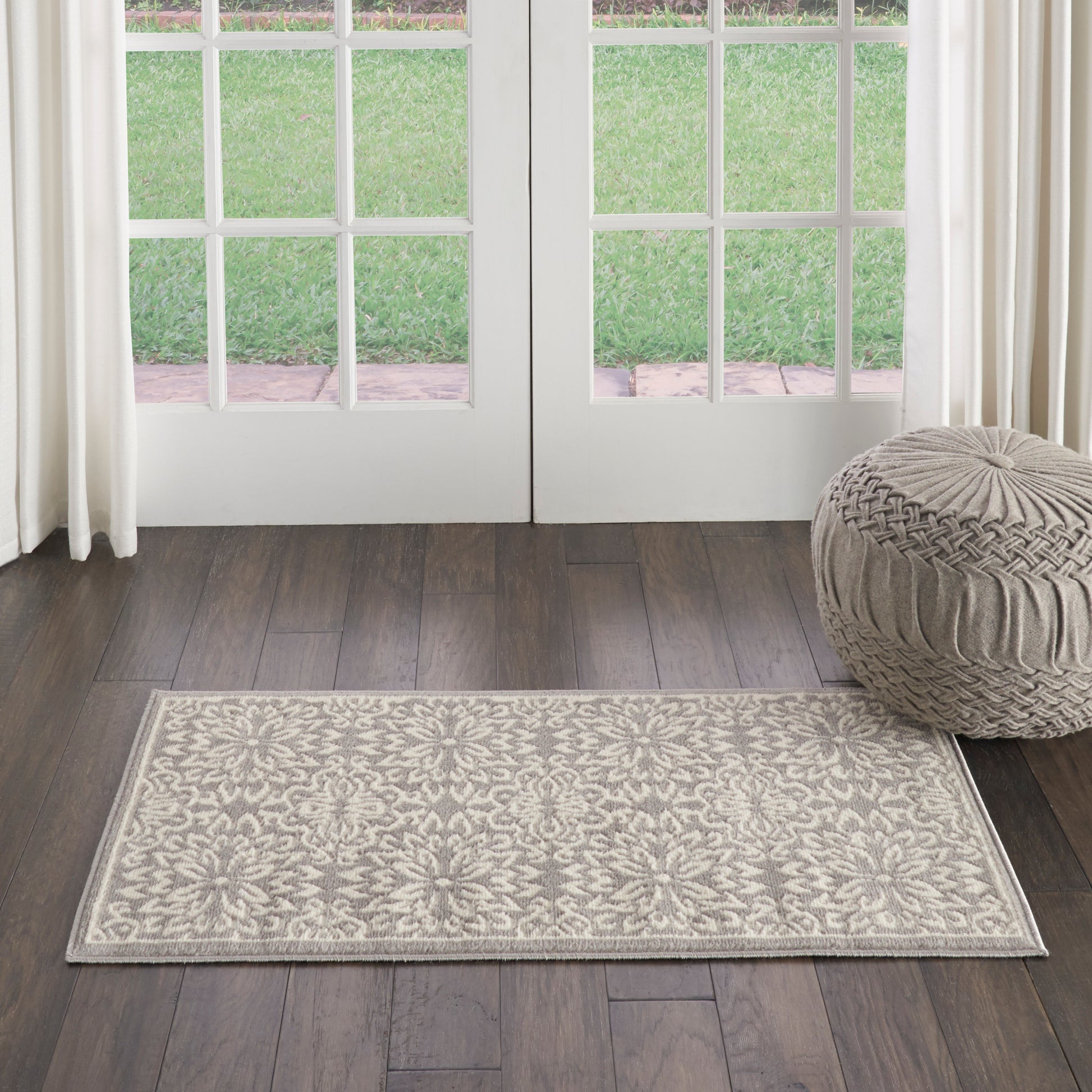Rectangular beige rug with ornate floral pattern on dark wood floor, beside a round knitted pouf in front of French doors.