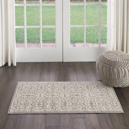 Rectangular beige rug with ornate floral pattern on dark wood floor, beside a round knitted pouf in front of French doors.