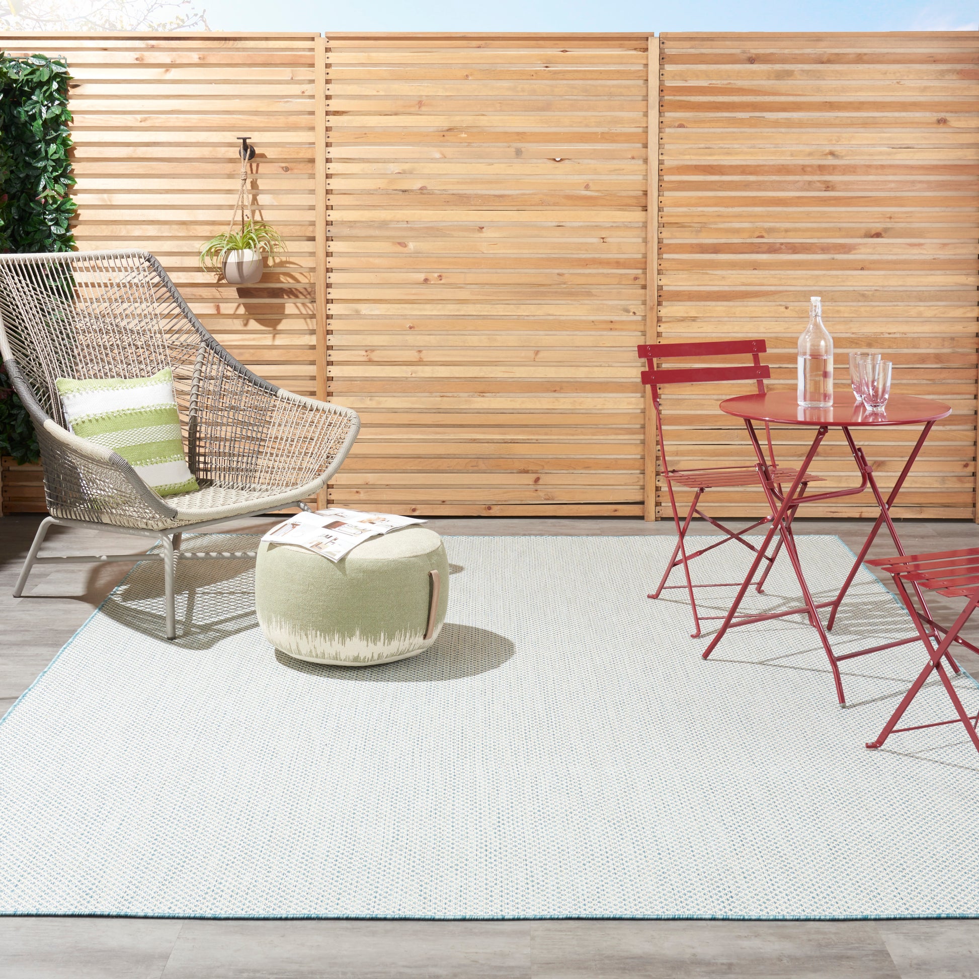 Outdoor patio with a wicker chair and green cushion, round pouf, light blue rug, and a red metal bistro table set.