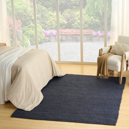 Navy blue textured woven area rug on light wood floor in a sunlit minimalist bedroom with beige bedding.