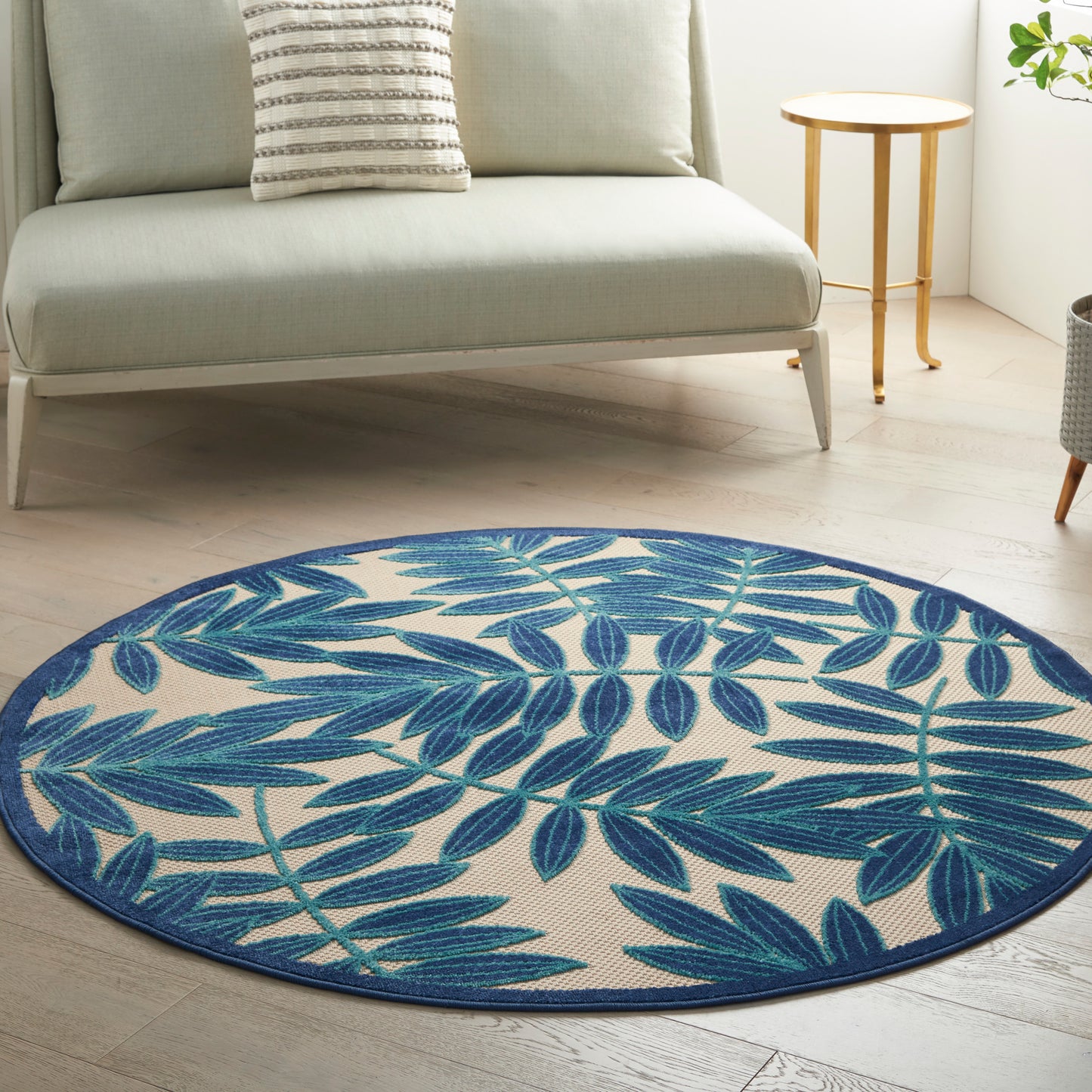 Round blue palm-leaf patterned rug on beige background in a light living room with a gray sofa and wooden side table.