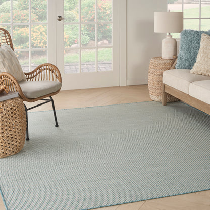 Light blue woven-area rug anchors a sunlit living room with rattan chairs, a beige sofa, and blue and cream pillows.
