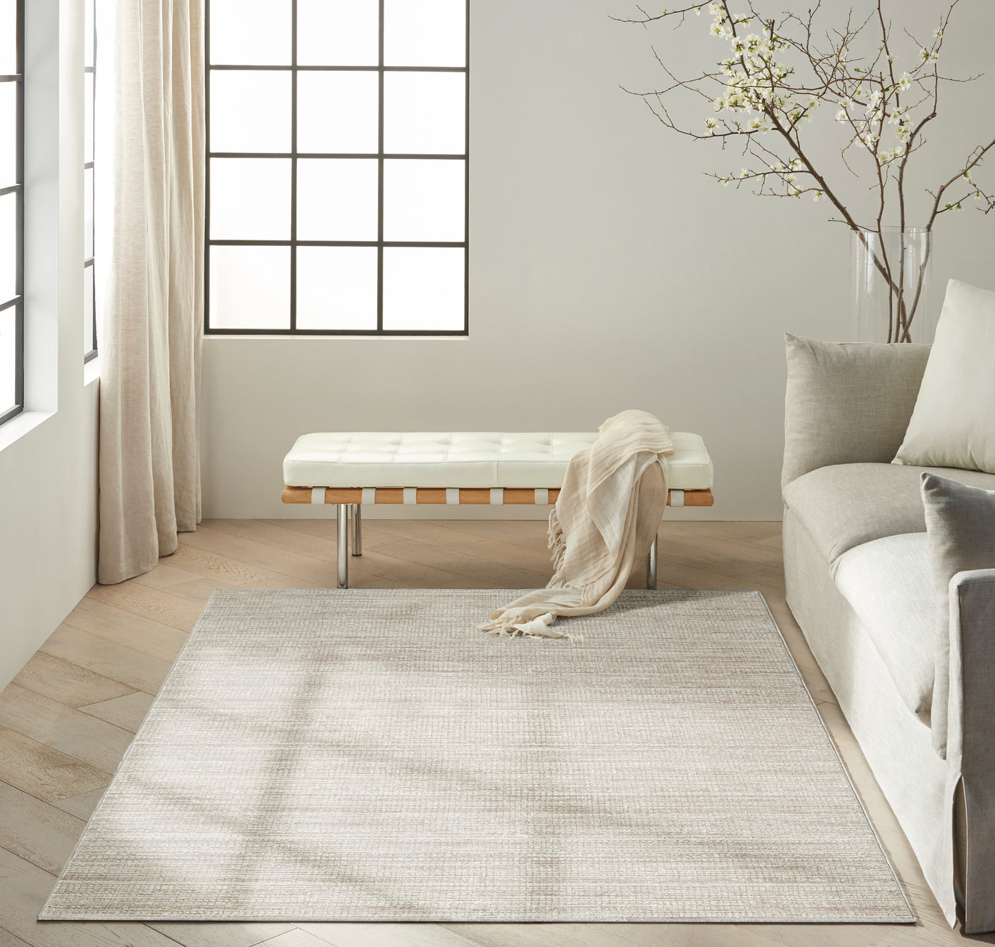 Minimalist living room with beige rug, white tufted bench with metal legs, wood floor, gray sofa, and tree branch decor.