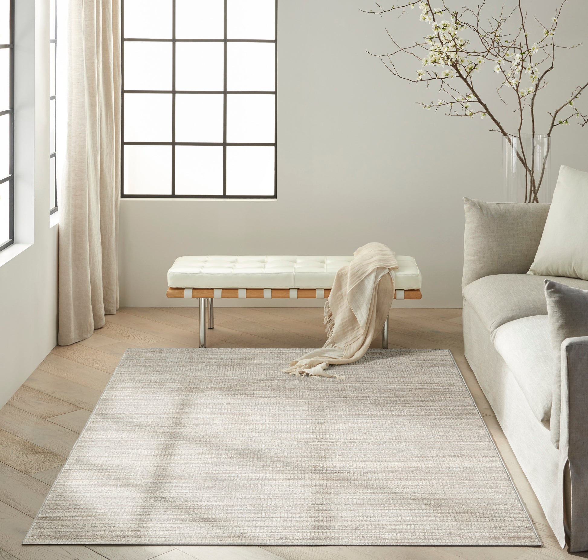 Minimalist living room with beige rug, white tufted bench with metal legs, wood floor, gray sofa, and tree branch decor.