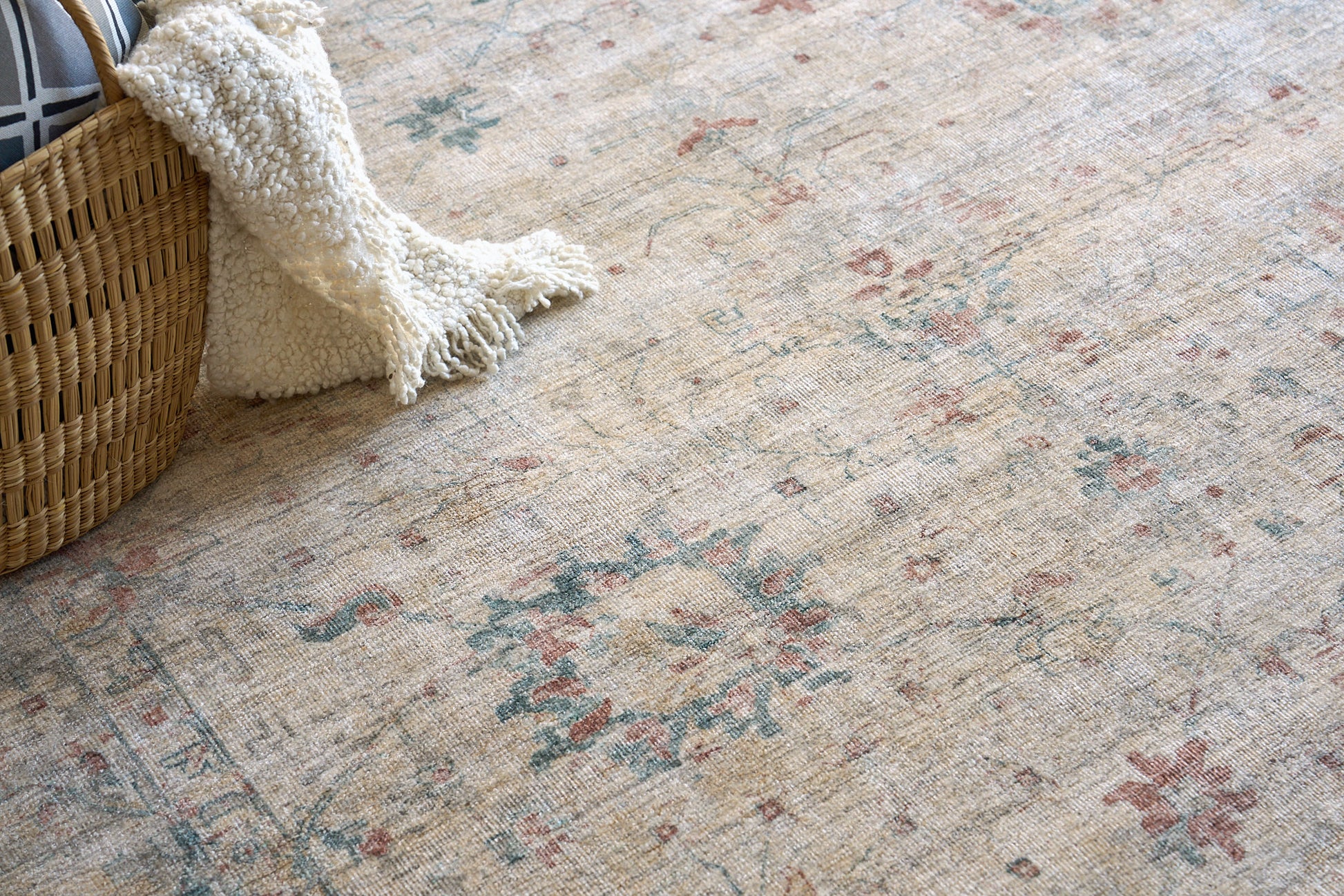 Beige floral rug with muted blue and red accents; woven basket with a cream plush throw.
