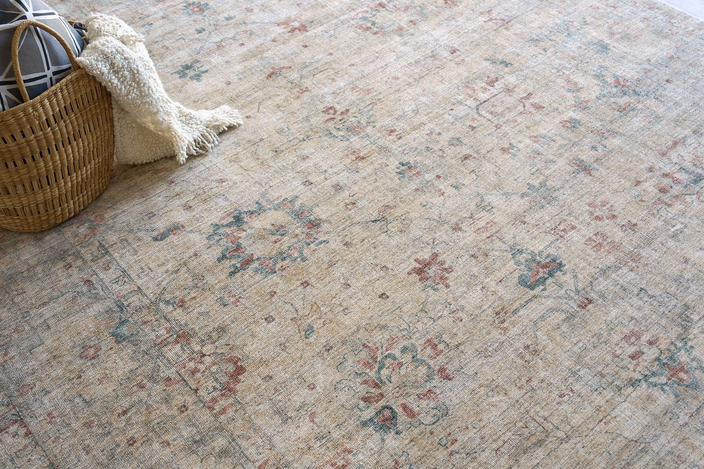 Beige vintage floral rug with faded blue and red motifs; wicker basket and cream textured throw nearby.