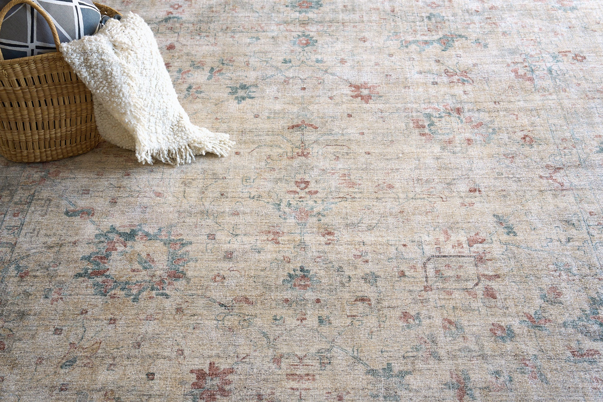 Beige oriental rug with muted blue and red floral pattern, cream textured throw draped over wicker basket.