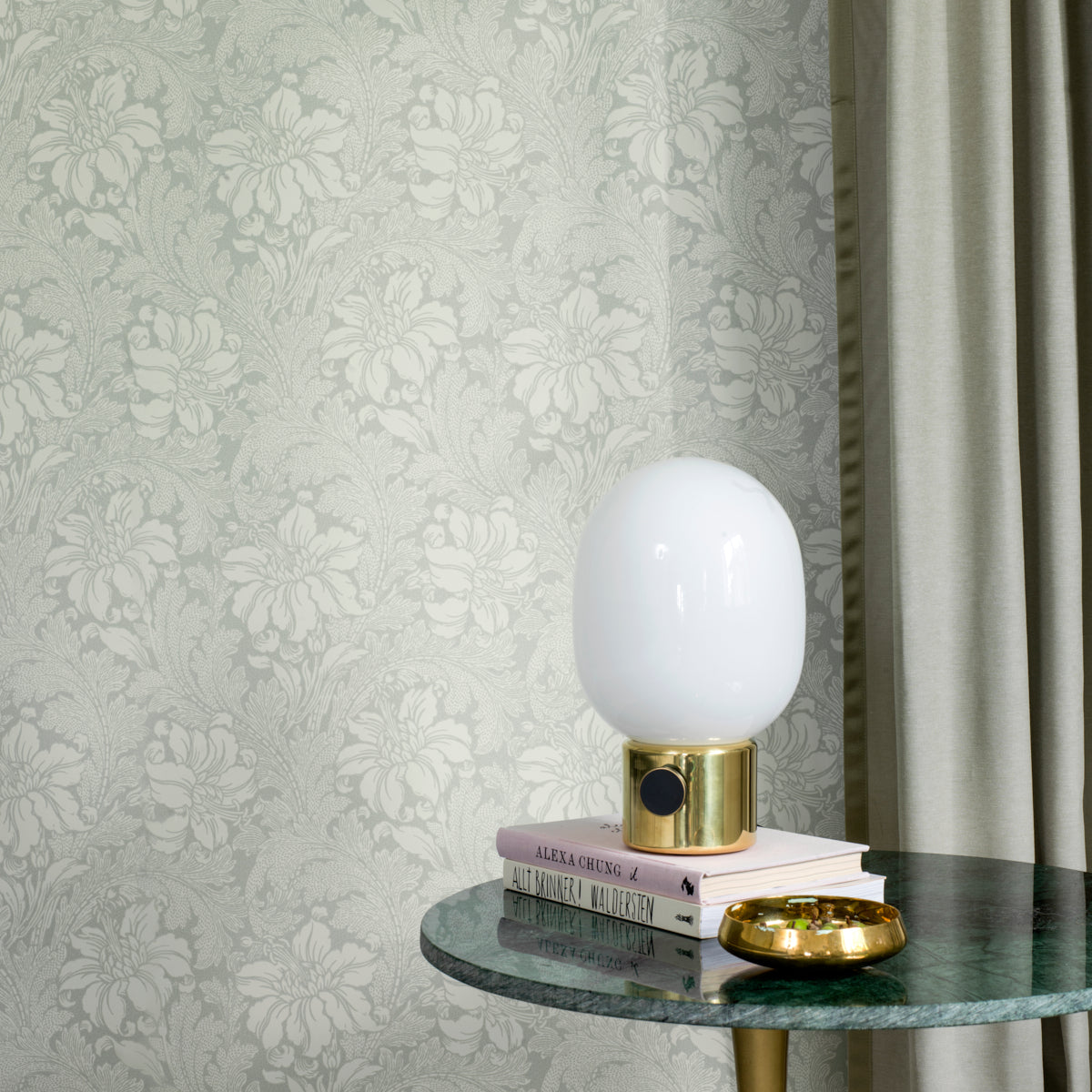 White glass table lamp with a gold base atop stacked books on a round green marble table, damask wallpaper backdrop.