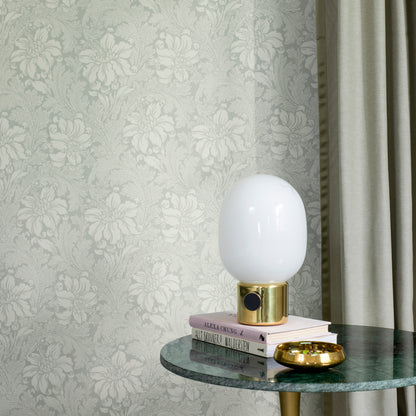 White glass table lamp with a gold base atop stacked books on a round green marble table, damask wallpaper backdrop.