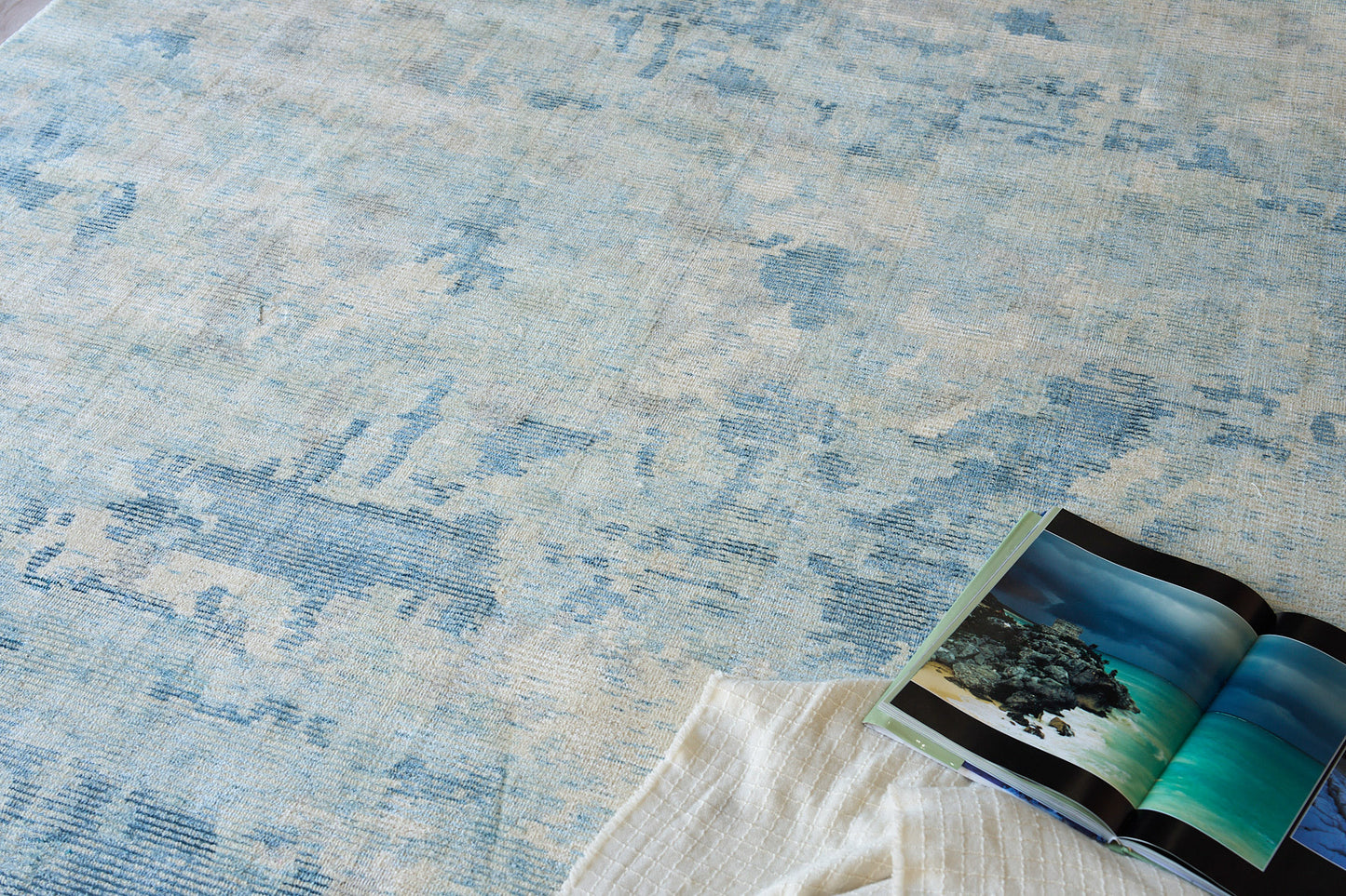 Blue & cream abstract patterned rug with a white quilted throw & an open travel magazine showing turquoise ocean photos.
