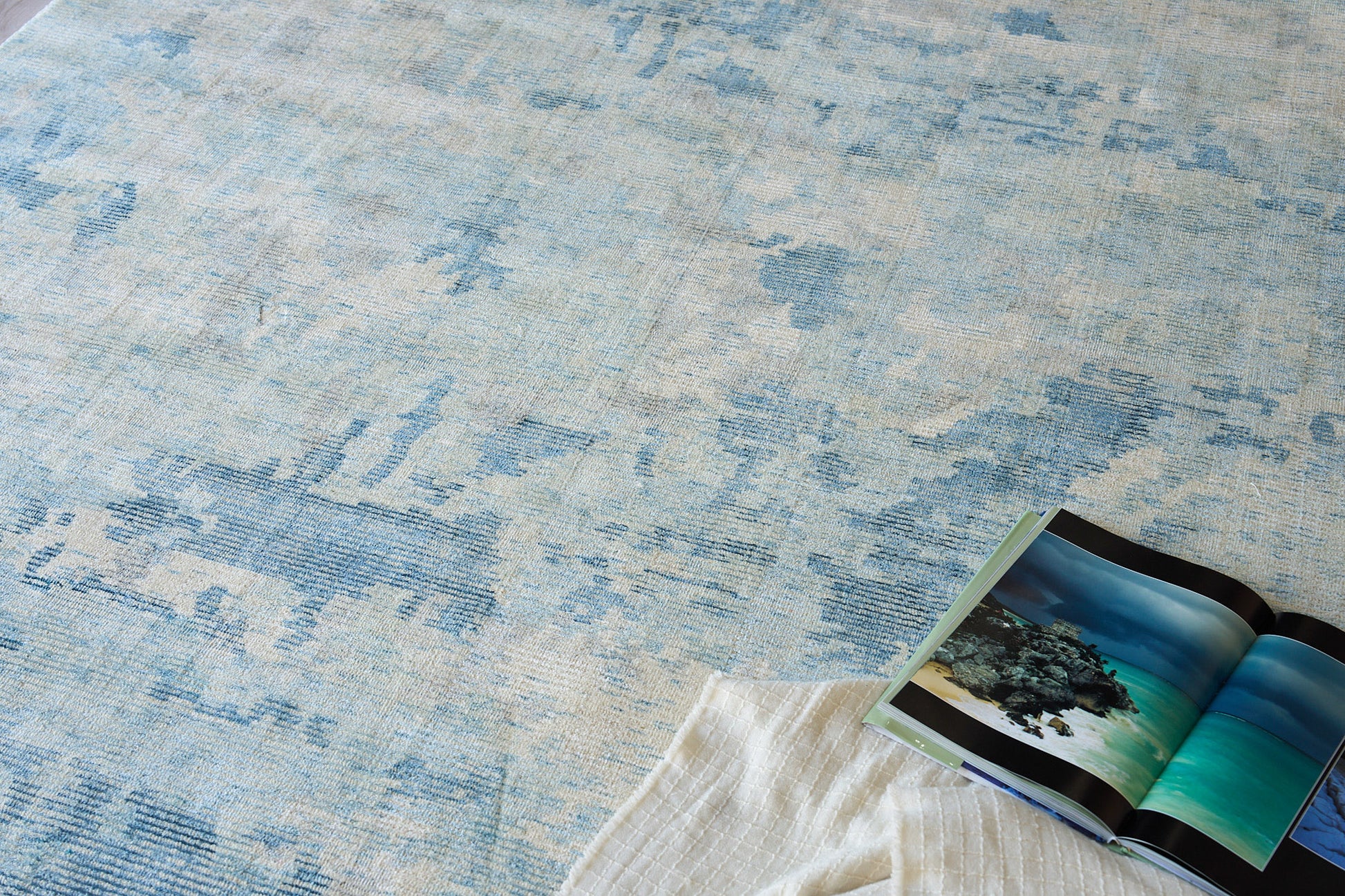 Blue & cream abstract patterned rug with a white quilted throw & an open travel magazine showing turquoise ocean photos.