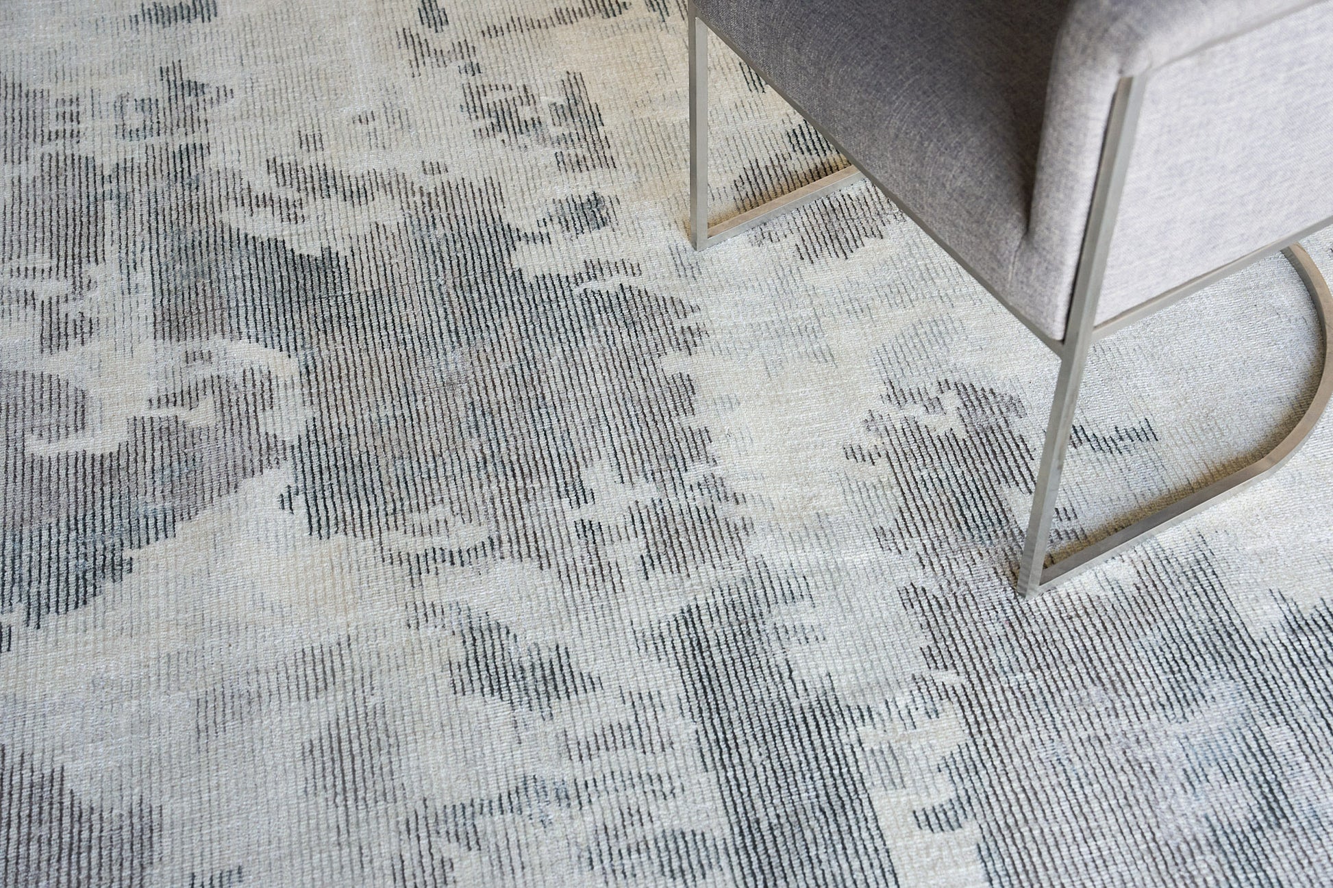 Soft textured area rug in gray-beige tones with abstract pattern; beside a gray upholstered chair with brass cantilever legs.