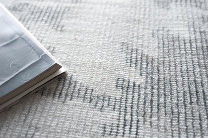 Gray rug with a textured surface and dark linear patterns; an open light gray magazine with white pages rests on it.