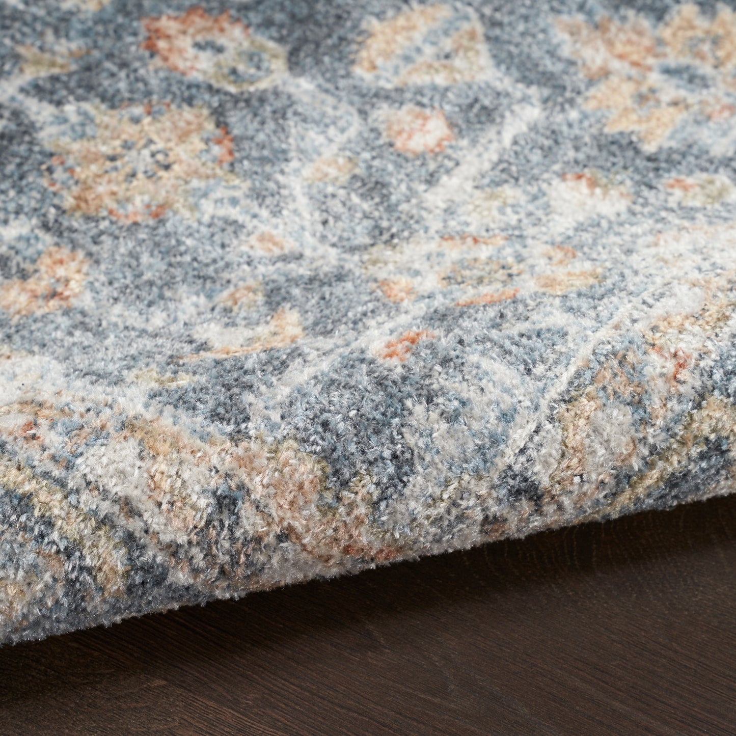 Close-up of a blue-gray floral rug with cream and peach accents, plush pile, resting on dark wood floor.