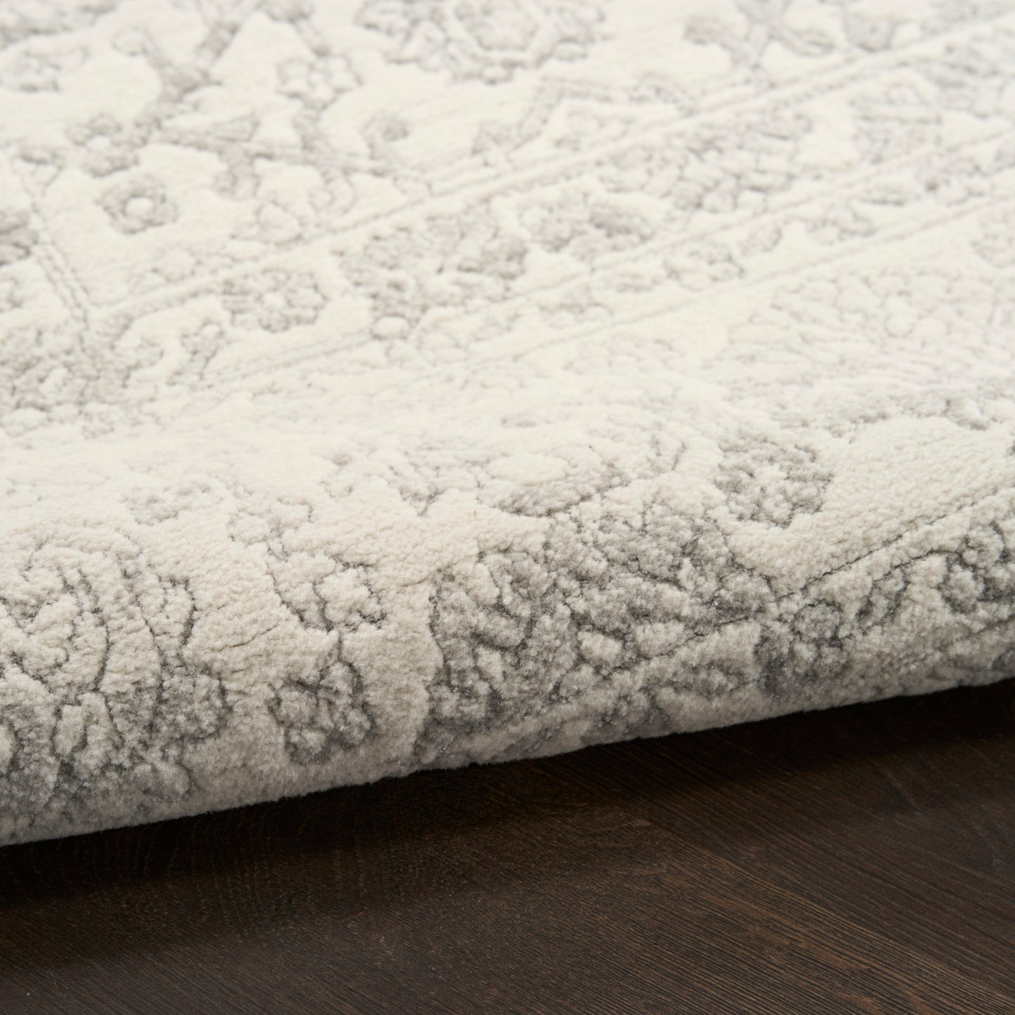 Light gray patterned rug with ornate floral motifs, folded edge on dark wood flooring.