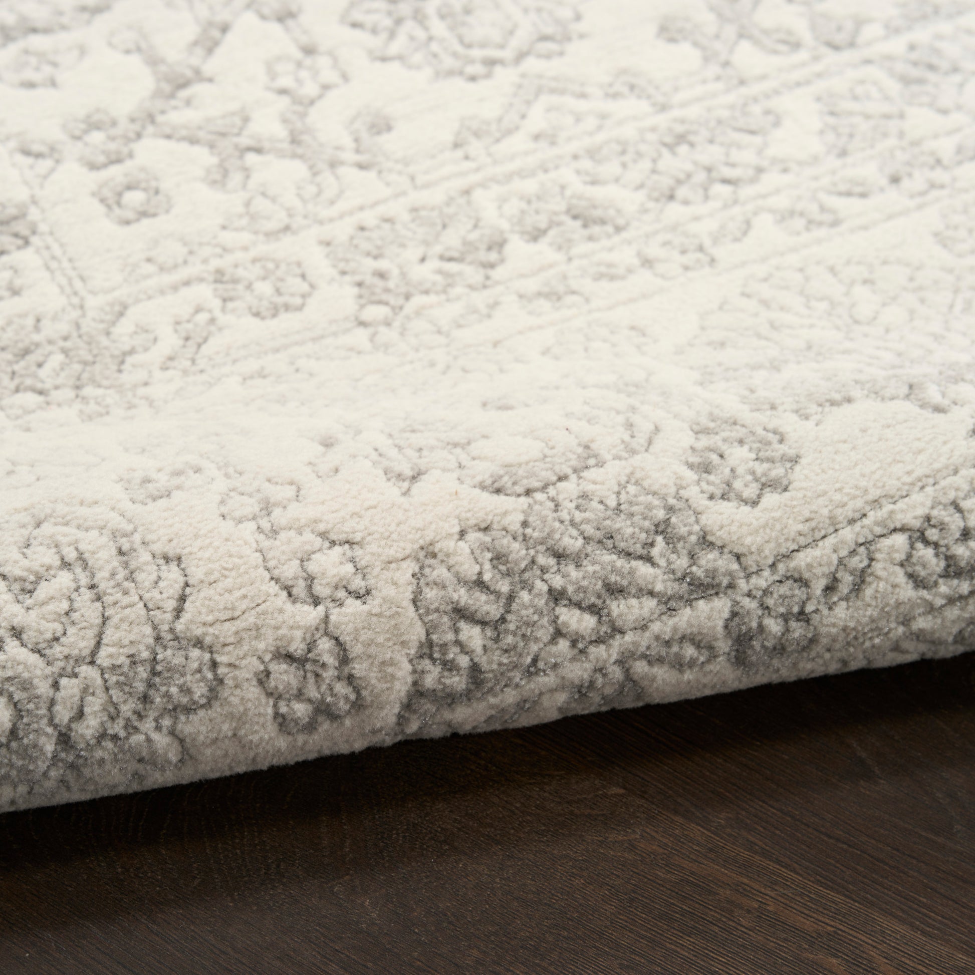 Light gray patterned rug with ornate floral motifs, folded edge on dark wood flooring.