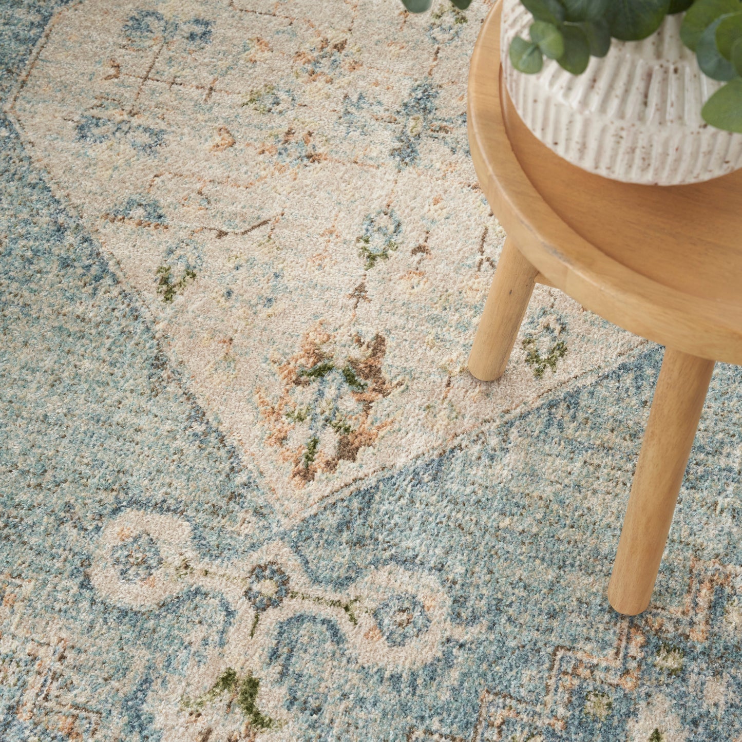 Blue and beige ornate round rug with floral motifs; light wood round side table with a white textured planter and greenery.