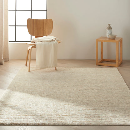 Natural wood chair with beige throw on a textured jute rug, beside a small wooden cube side table with vase and bowl in a bright minimalist room.