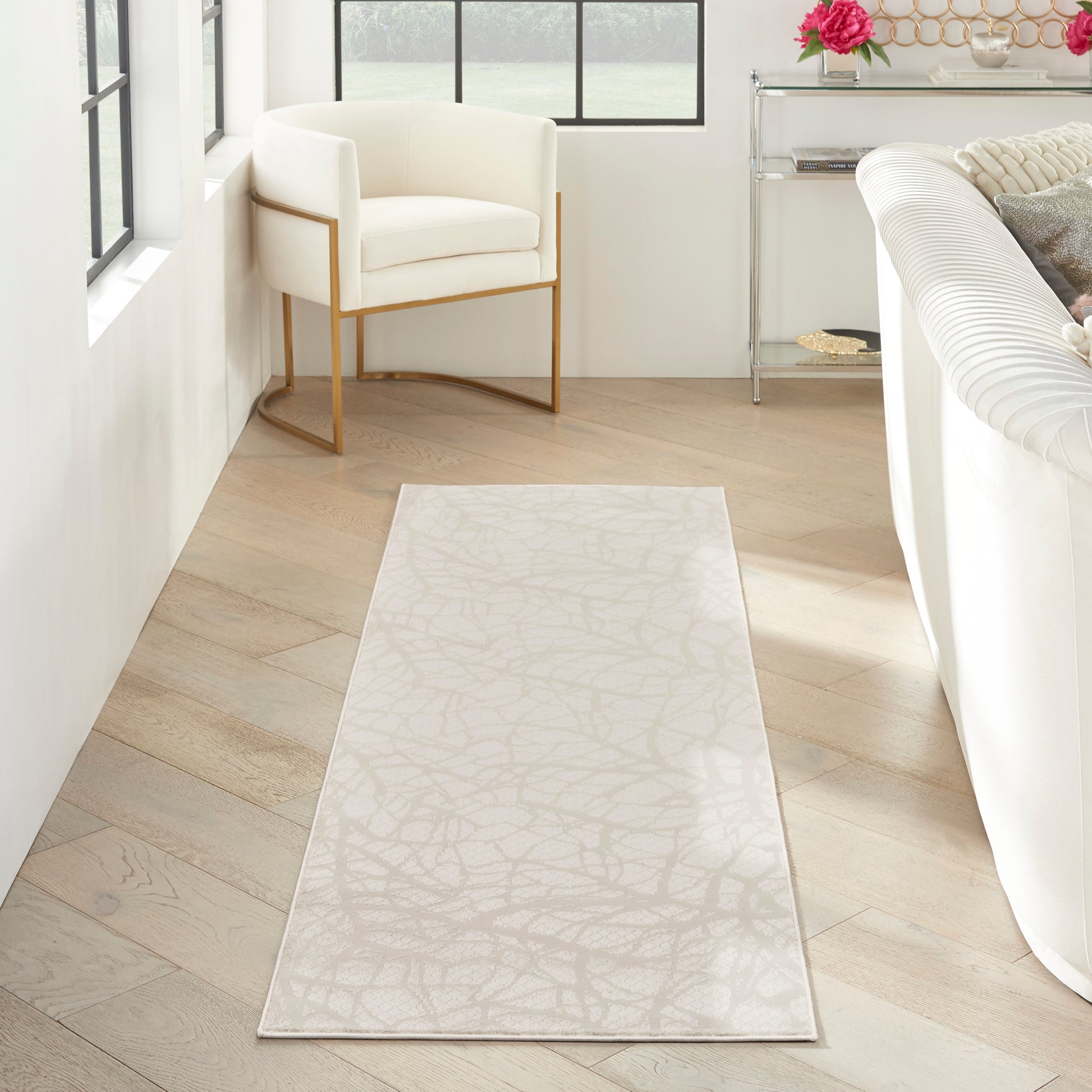 Long cream runner rug with subtle abstract leaf pattern on light wood floor beside white sofa and gold-framed chair.