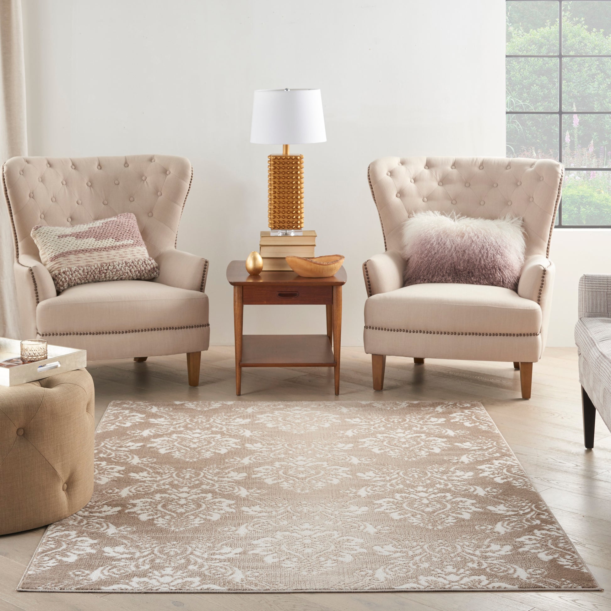 Two beige tufted armchairs with nailhead trim flank a wooden end table and lamp in a cozy beige living room with a rug.