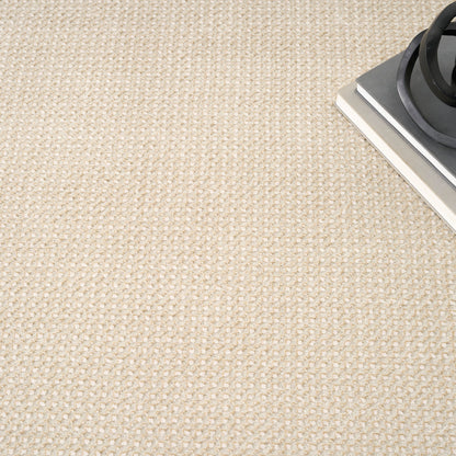 Beige woven rug with loop texture; corner shows a metal panel and black circular burner ring.