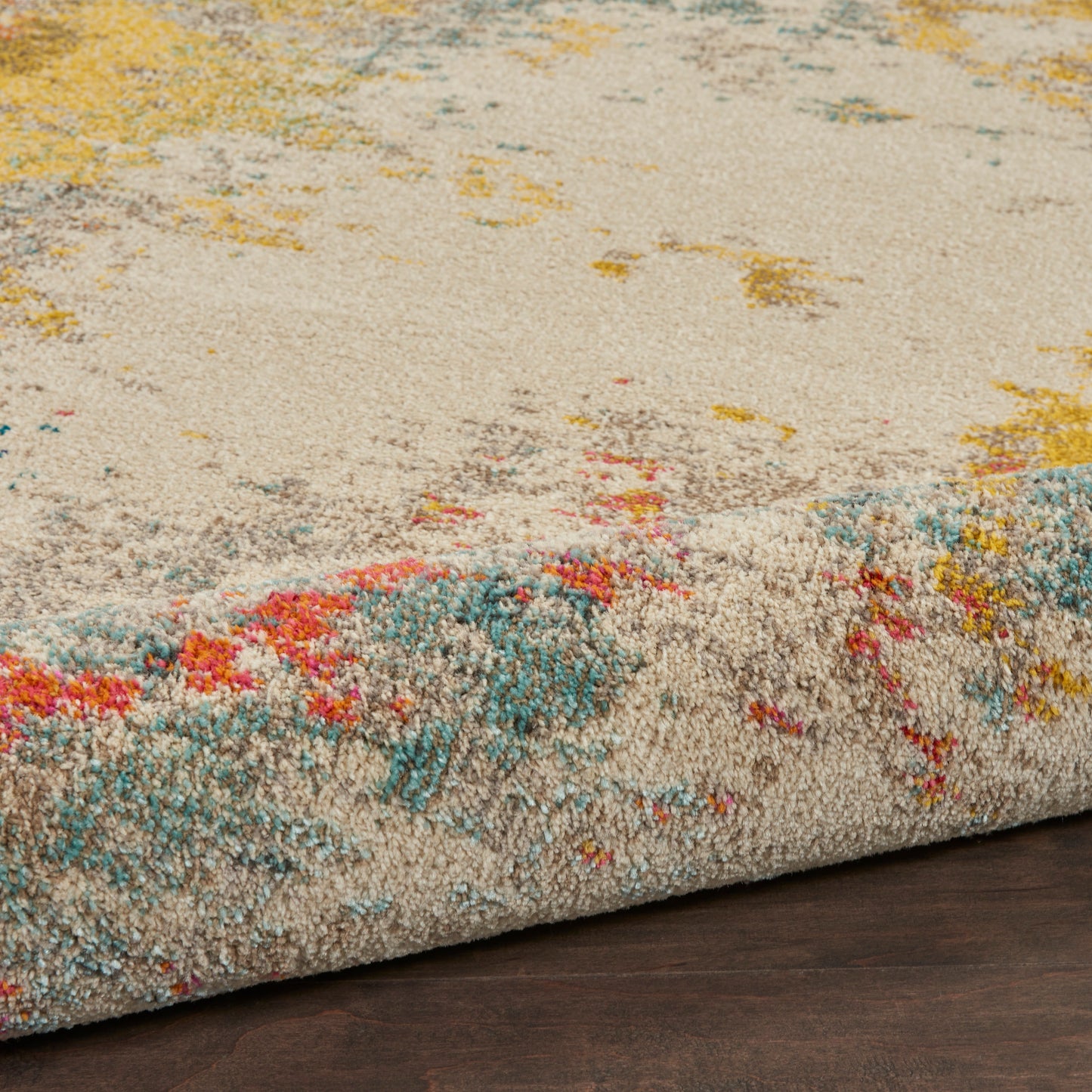 Beige plush rug with abstract multicolor accents in teal, yellow, pink and orange; rolled edge on dark wood floor.