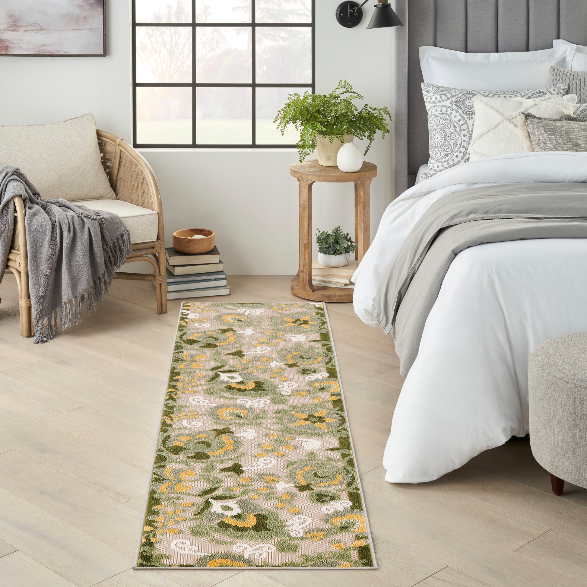 Long botanical flatweave rug runner in greens, yellows and ivory on a light wood floor beside a white bed in a modern bedroom