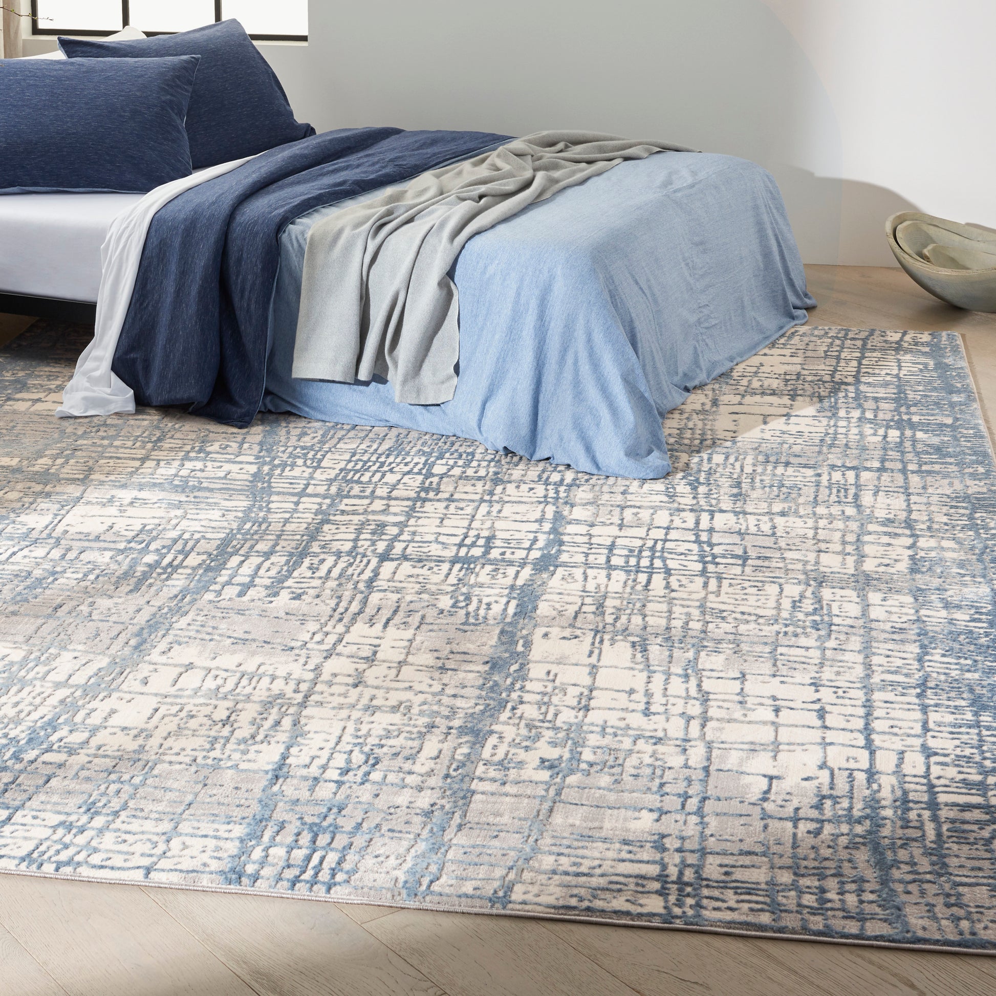 Bed with light blue duvet, navy throw, gray blanket and white sheet on a blue-gray abstract patterned rug.