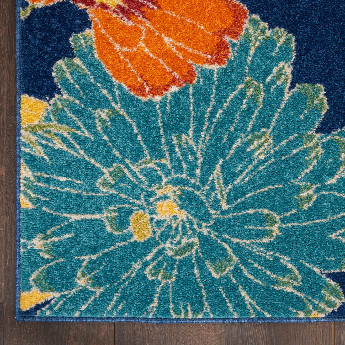 Tufted floral rug with teal petals and an orange bloom, yellow accents on a dark blue background, laid on a wooden floor.