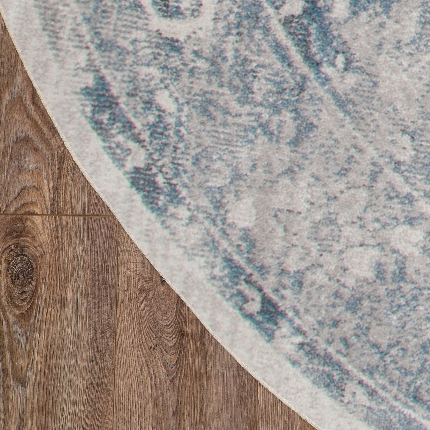 4' round blue-gray patterned area rug partially over brown wood floor, showing curved edge & textured design.