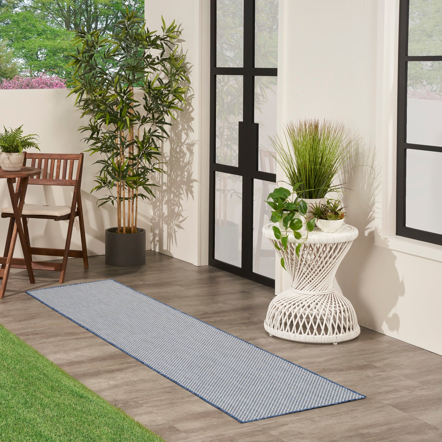 Blue striped outdoor runner rug on wood floor beside black-framed glass doors, with potted plants and wicker furniture.