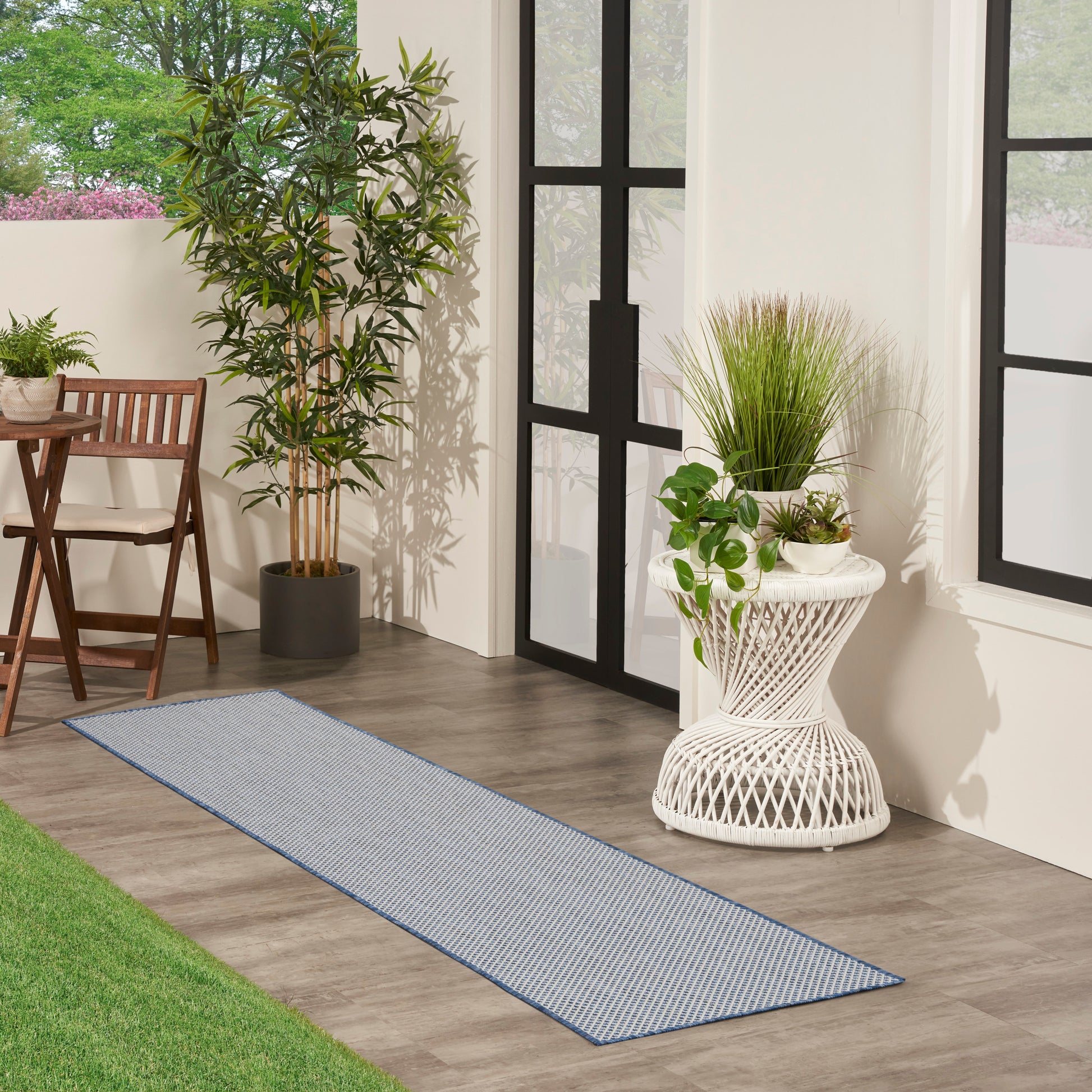 Blue striped outdoor runner rug on wood floor beside black-framed glass doors, with potted plants and wicker furniture.