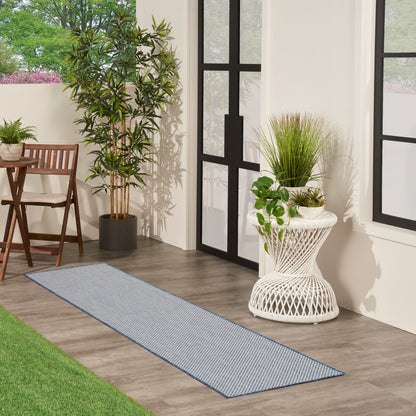 Blue striped outdoor runner rug on wood floor beside black-framed glass doors, with potted plants and wicker furniture.