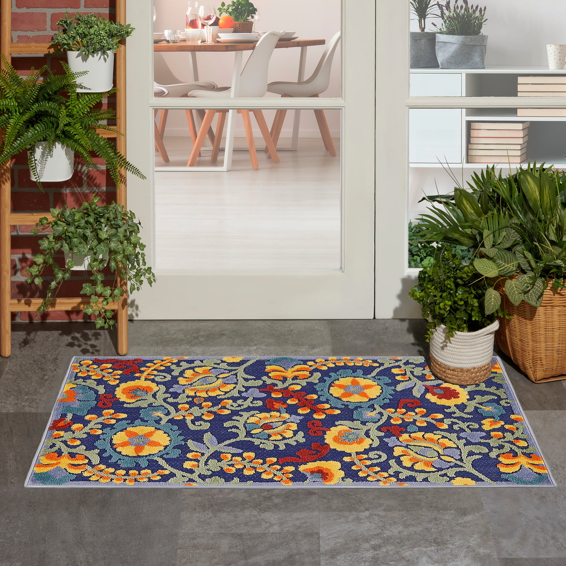 Colorful floral outdoor rug on grey entry tiles, blue background with yellow and orange blooms, beside potted plants.