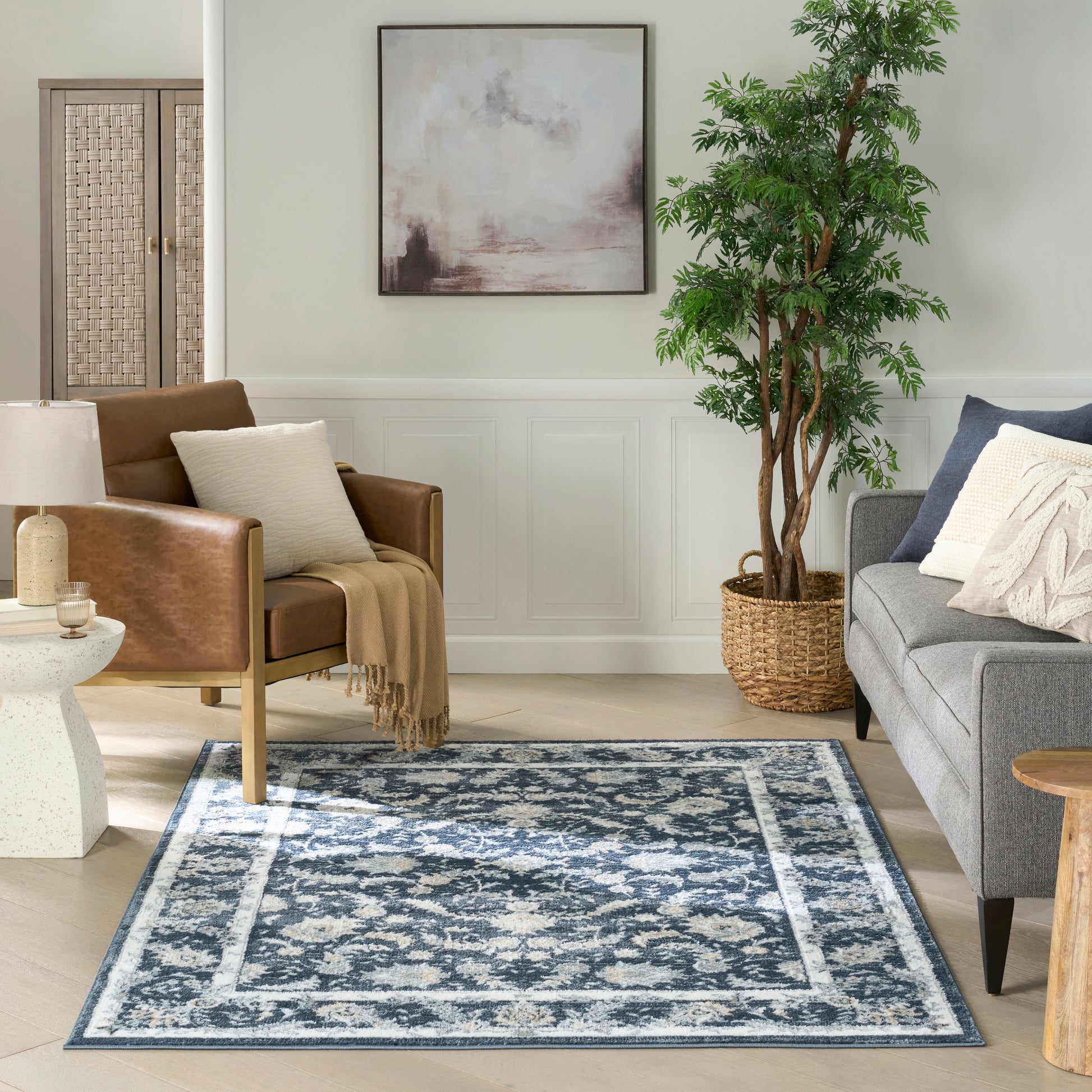 Blue patterned area rug on light wood floor with gray sofa, tan leather chair, cream throw pillows, and tall potted plant near abstract wall art.