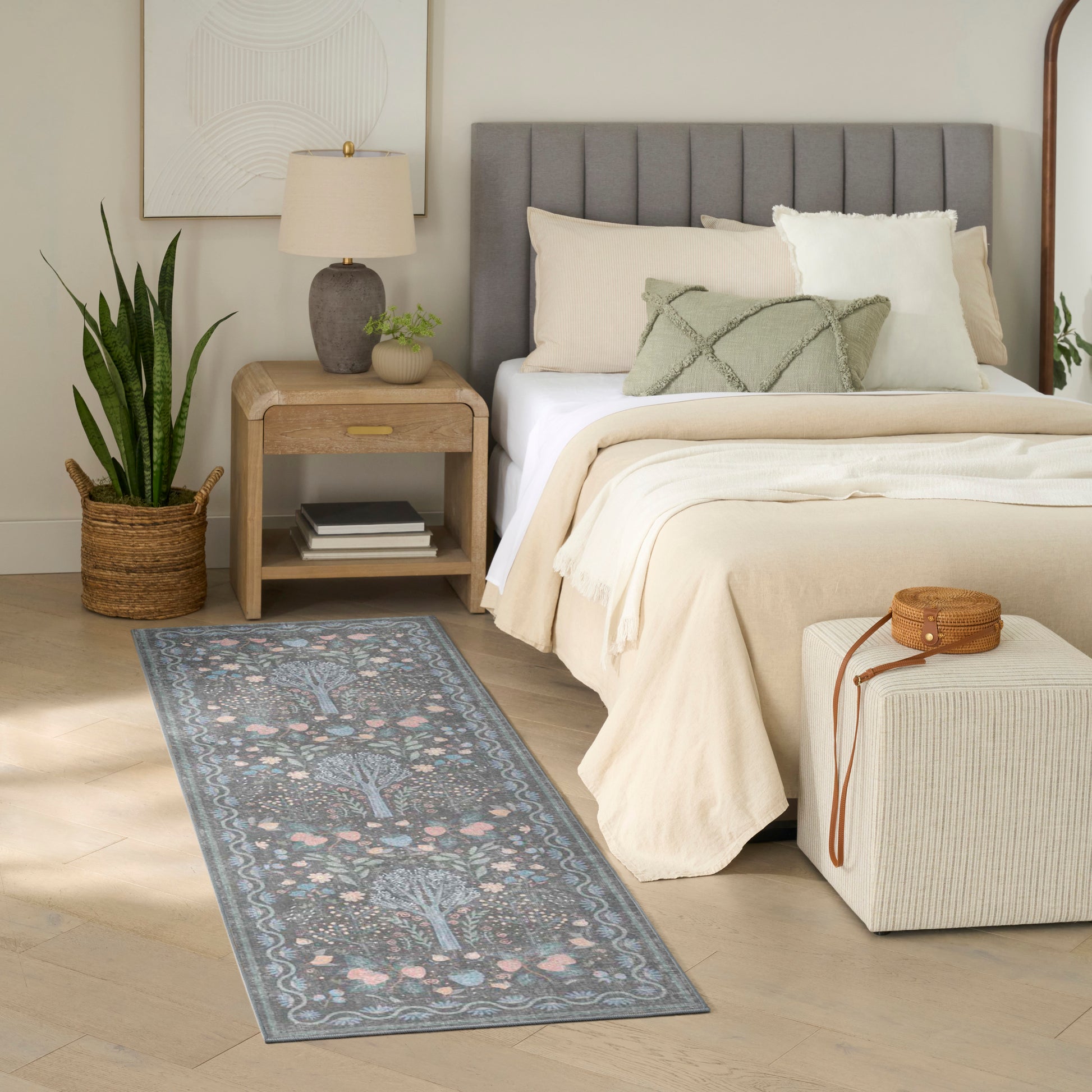 Beige bedroom with a tufted gray headboard, cream bedding, blue floral runner rug, and wooden nightstand with a potted plant.