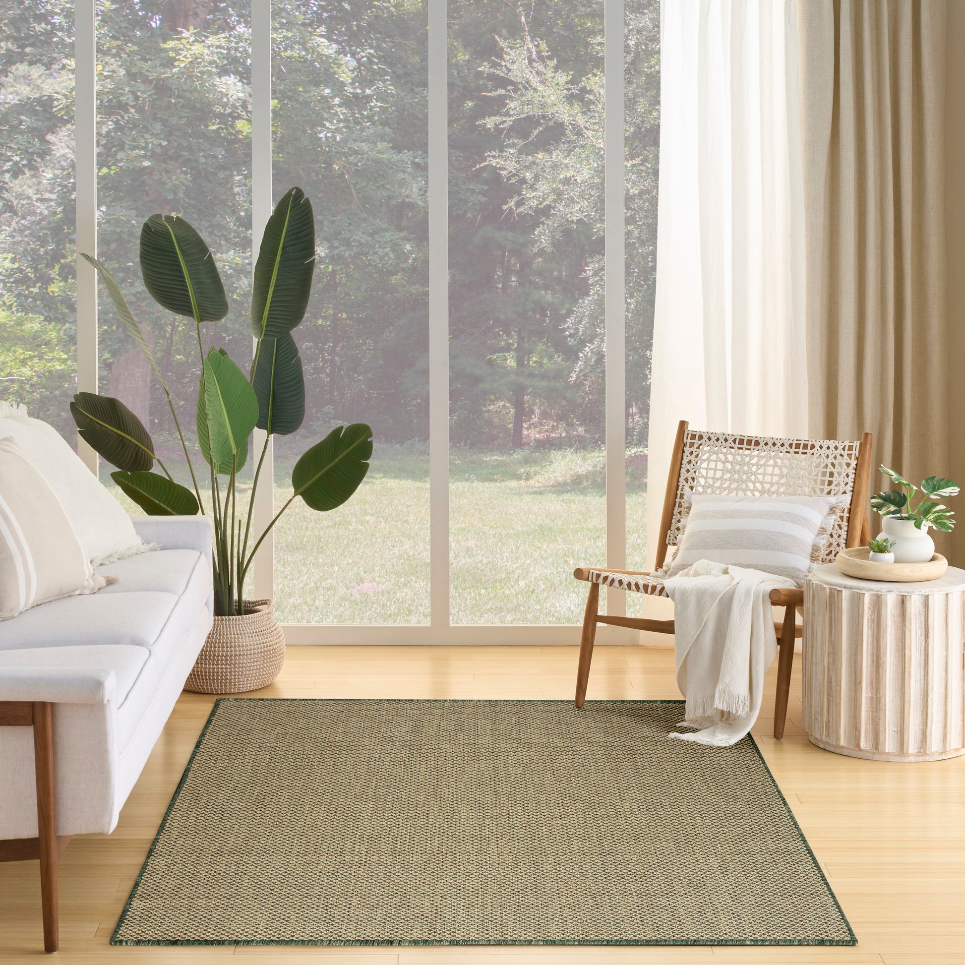 Living room scene with olive area rug, light wood floor, white sofa, woven chair, tall plant, and large windows.