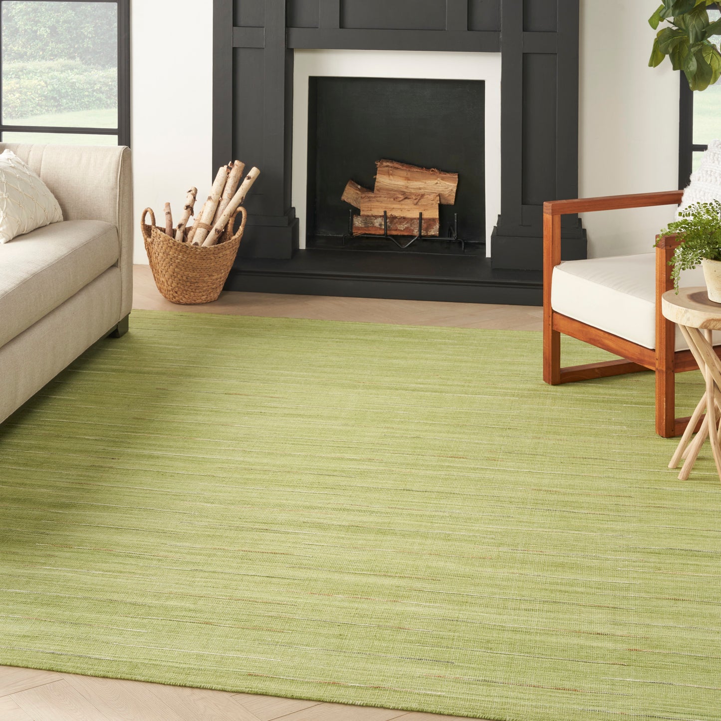 Light-green area rug (9' x 12') in a living room with a black fireplace, log basket, beige sofa & wooden armchair.