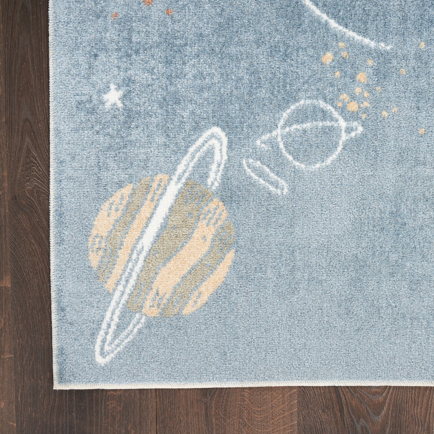 Blue space-themed rug with a beige ringed planet, white orbital rings and tiny stars on a wooden floor.
