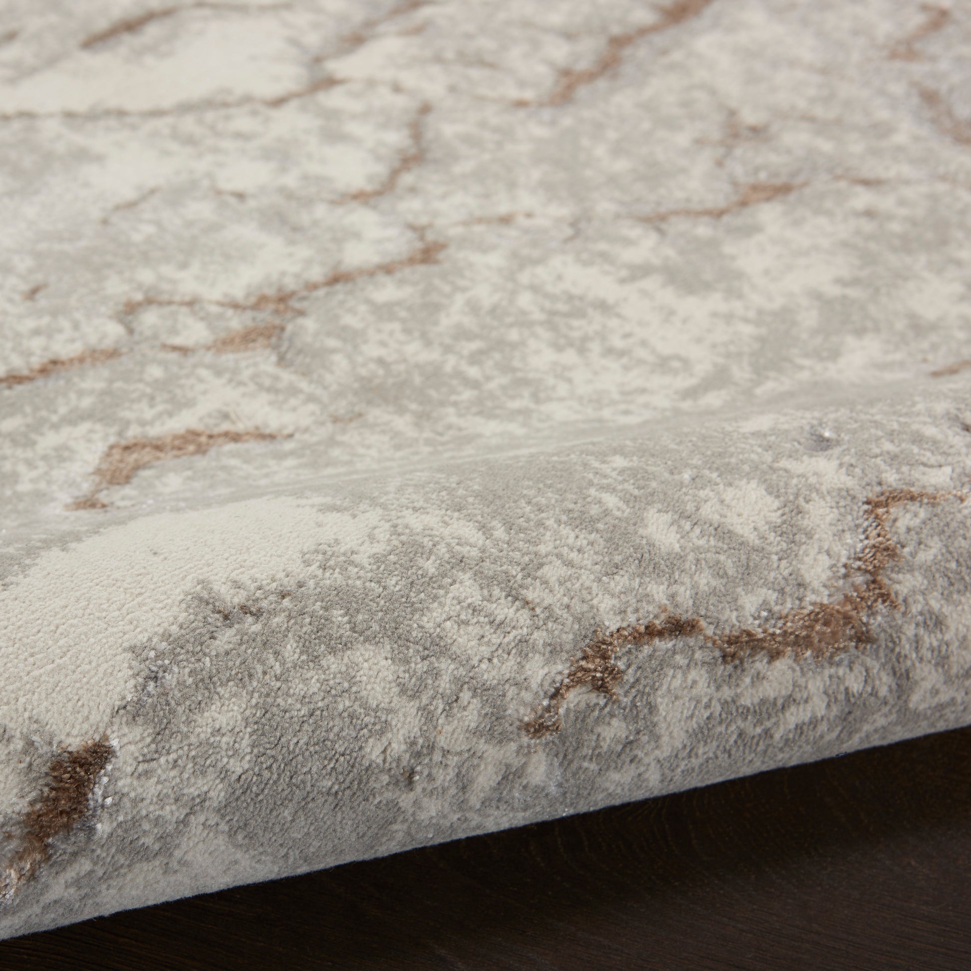 Close-up of a light grey rug with brown abstract lines, plush cut-pile texture, folded edge on dark wood floor.