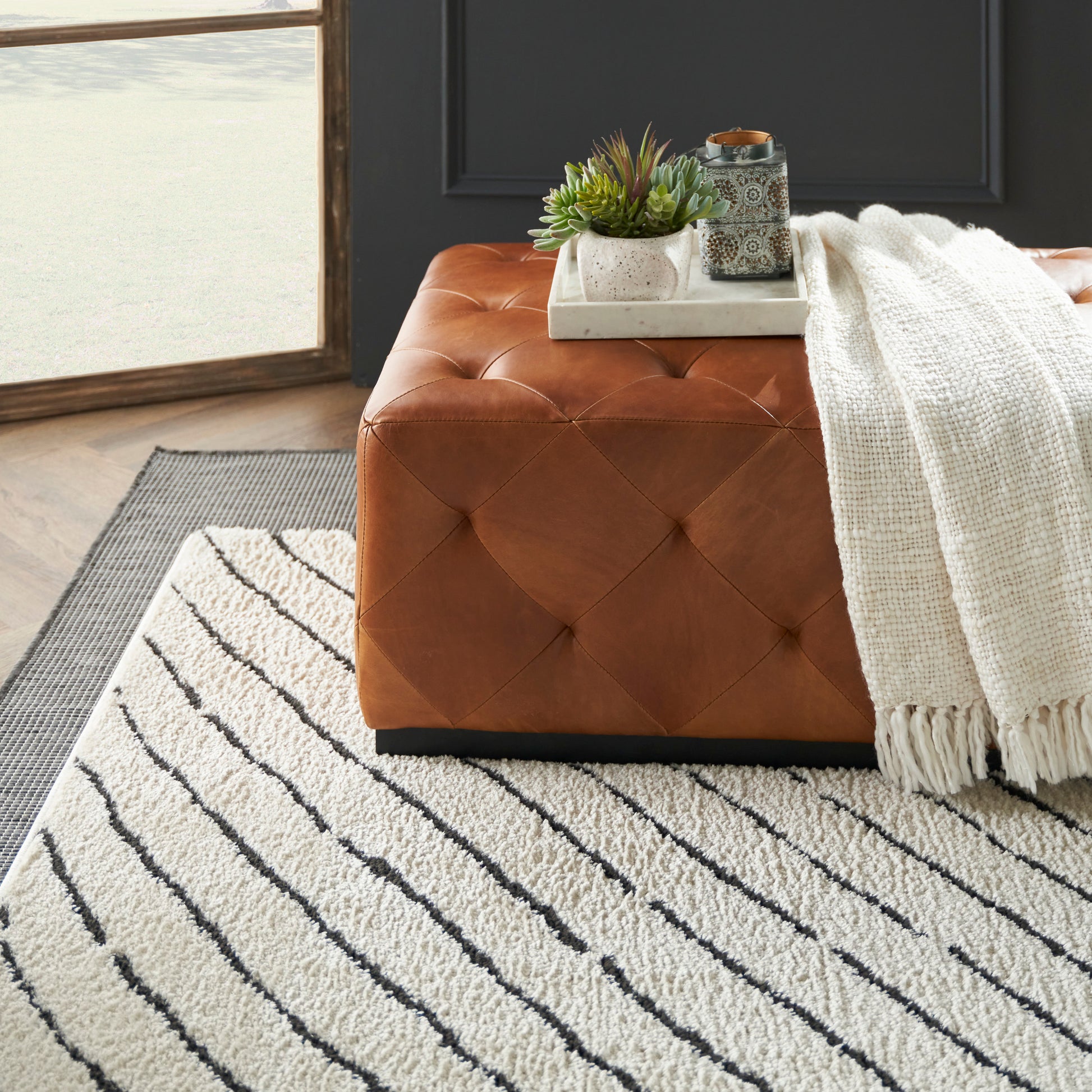 Brown tufted leather ottoman with diamond stitching on a striped cream rug; white knit throw draped, tray with succulents on top.