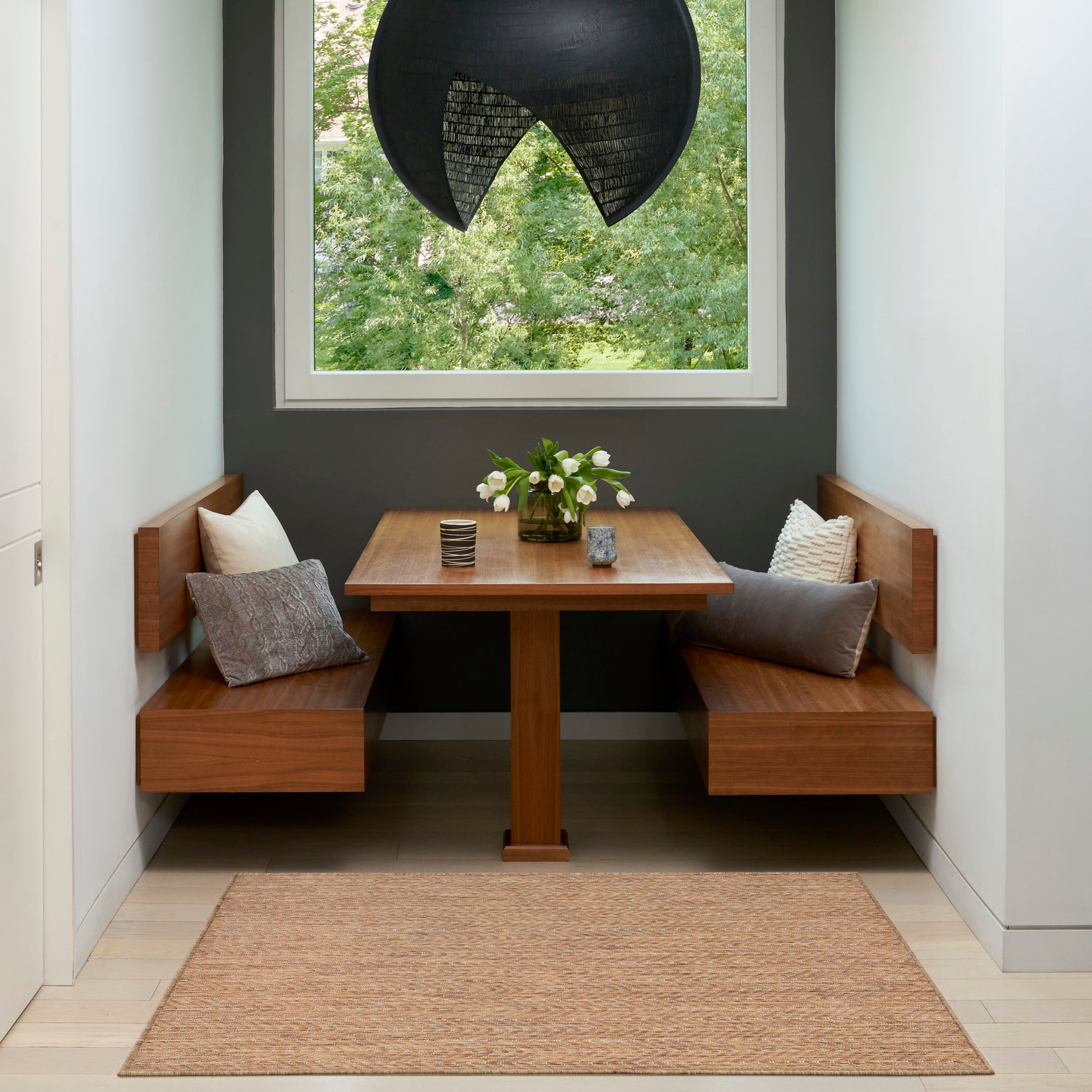Cozy wooden dining nook with built-in benches, square table, cushions, black pendant lamp, window greenery, beige rug.