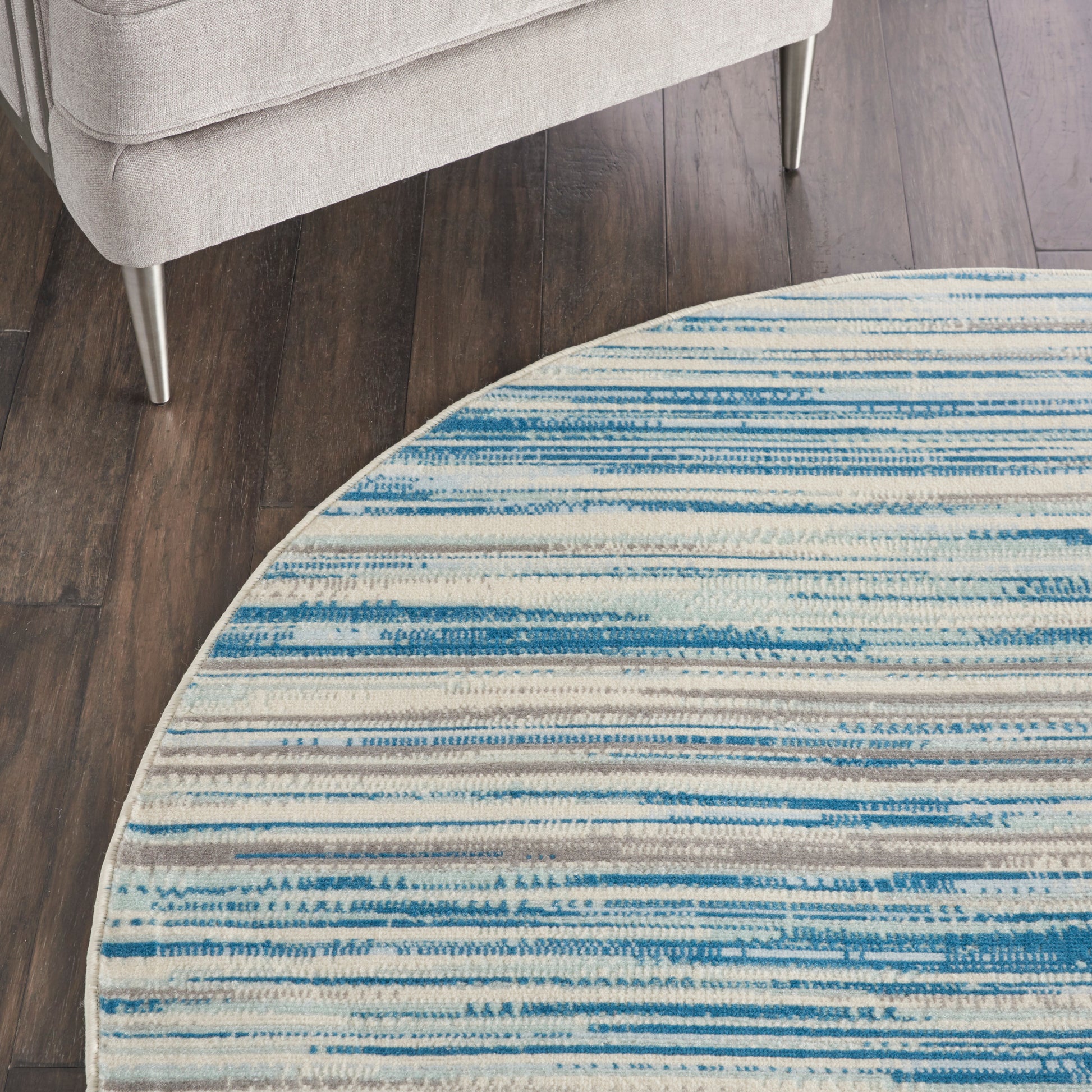 Round rug with blue striped pattern on white, on dark wood floor beside a light gray sofa with metal legs.