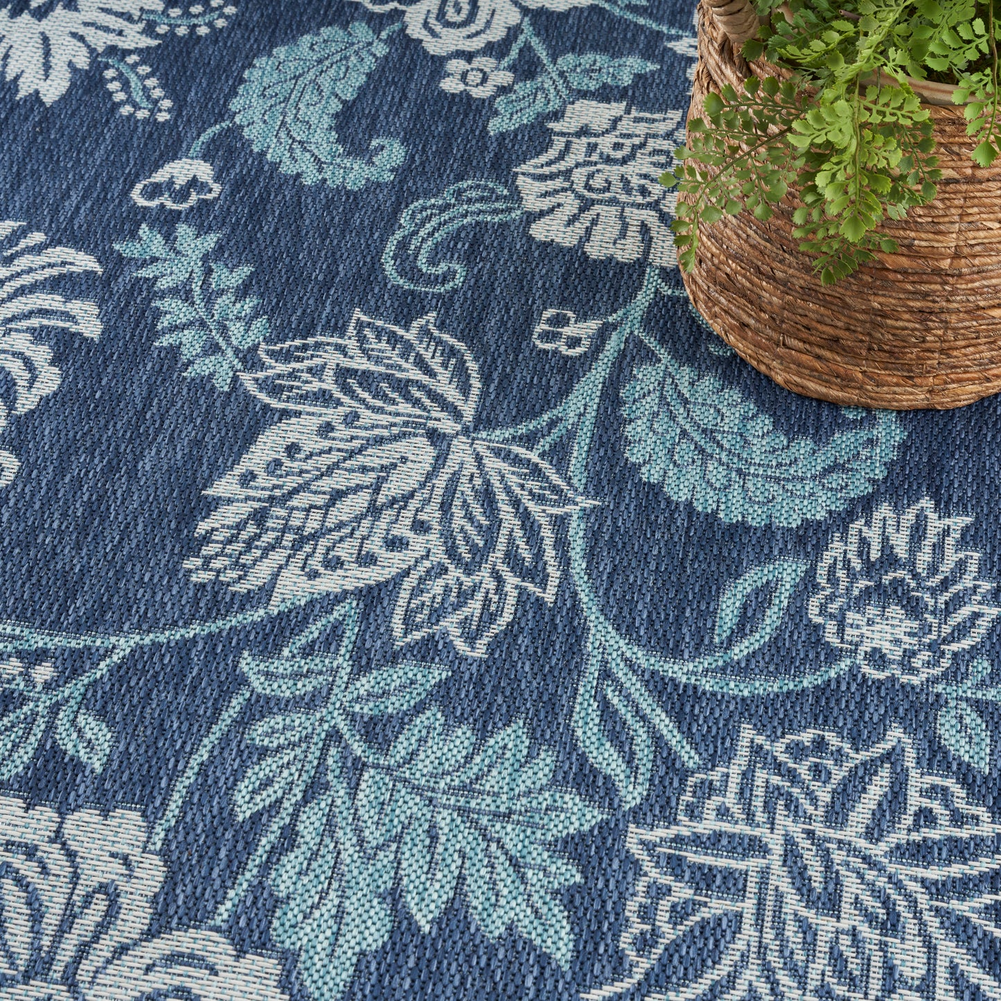 Dark blue woven rug with teal and white floral damask pattern; corner wicker basket with green plant.