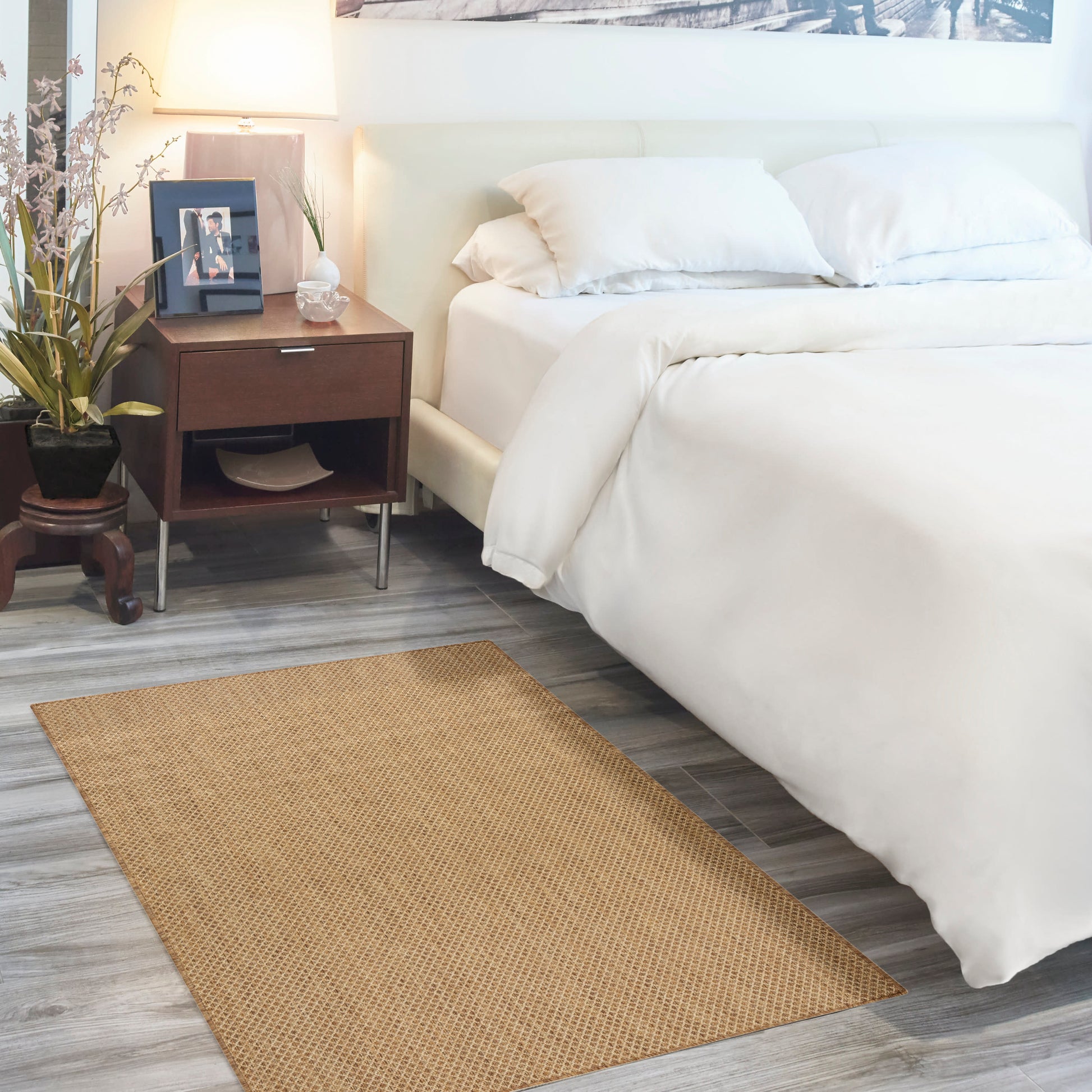 White bed with plush duvet and pillows, dark wood nightstand, lamp, plant, and beige woven rug on gray wood floor.