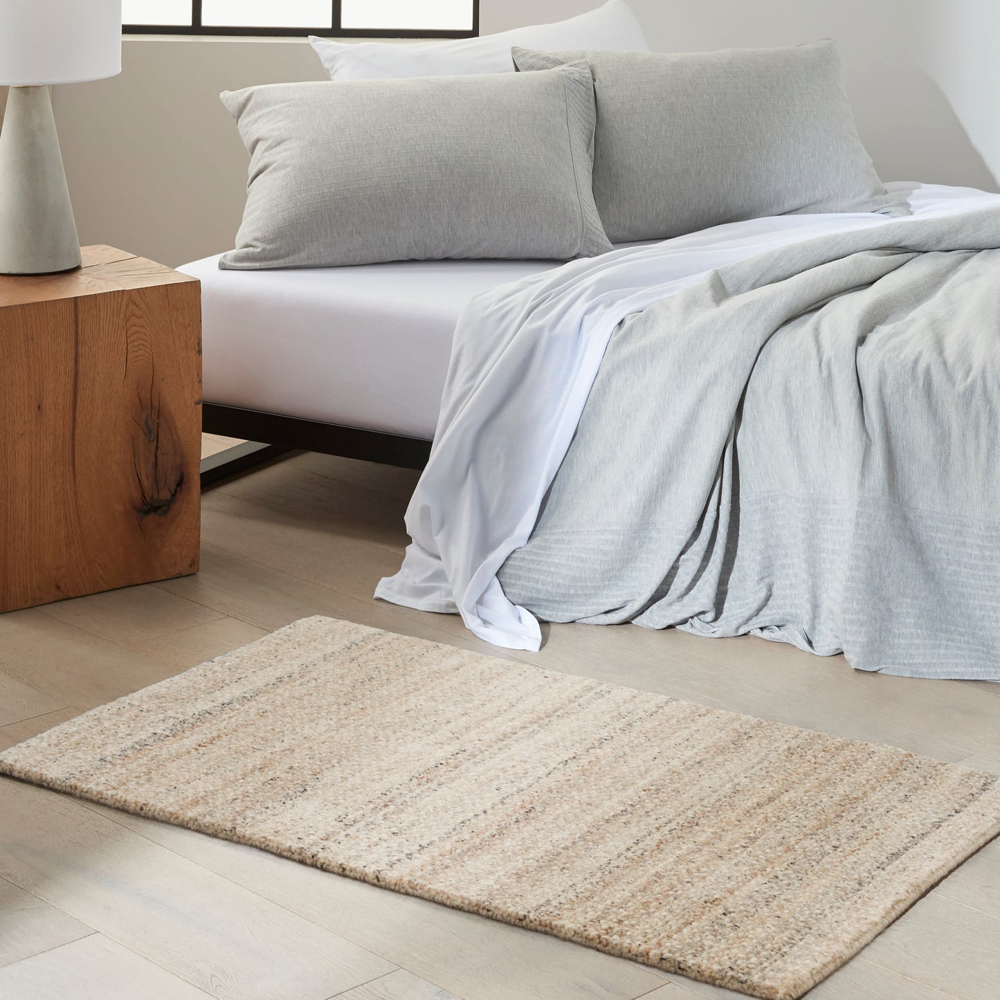 Minimalist bedroom with a low platform bed, light gray bedding, beige woven rug, and a wooden block nightstand with lamp.