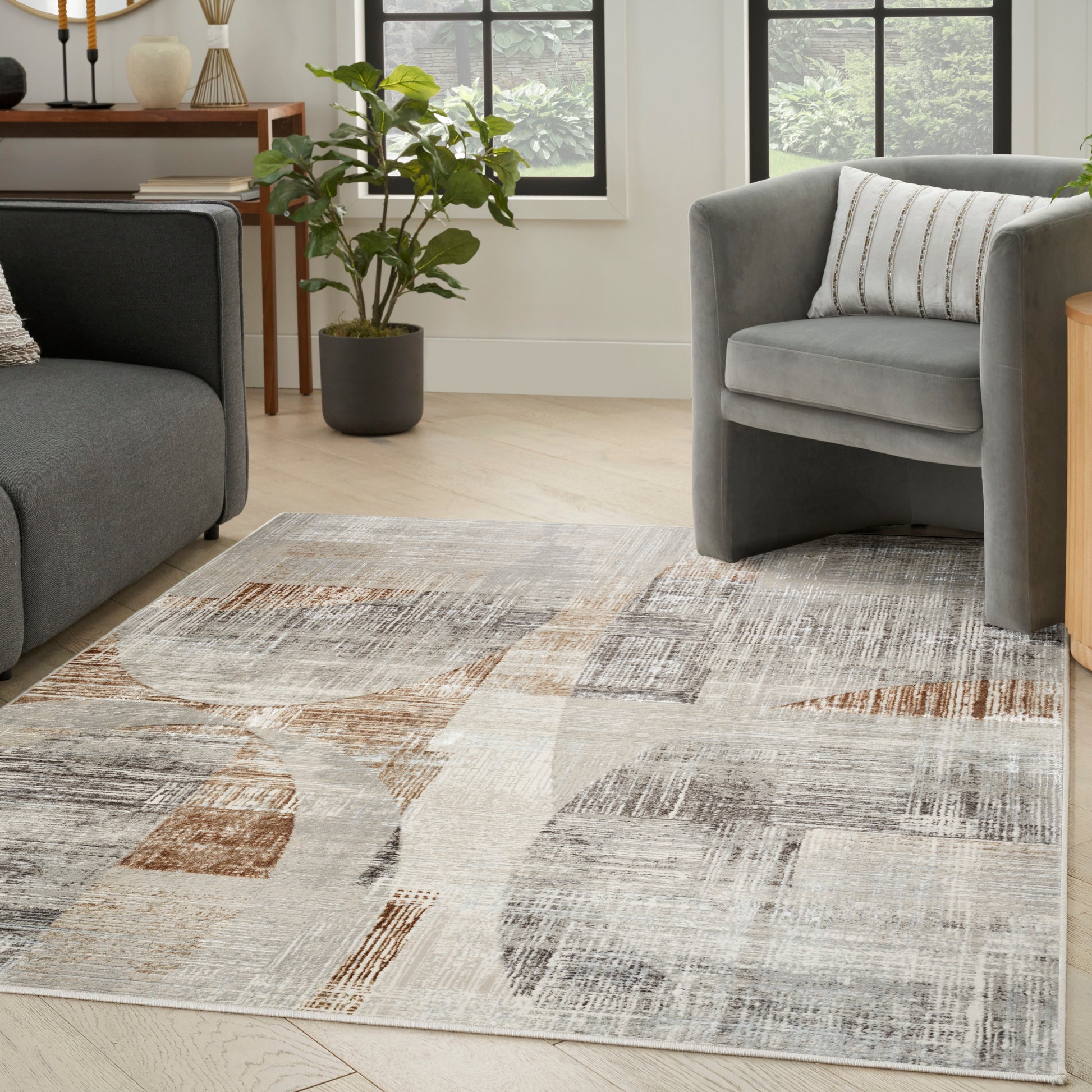 Geometric abstract area rug in beige, gray and taupe on light wood floor beside a gray sofa and armchair.