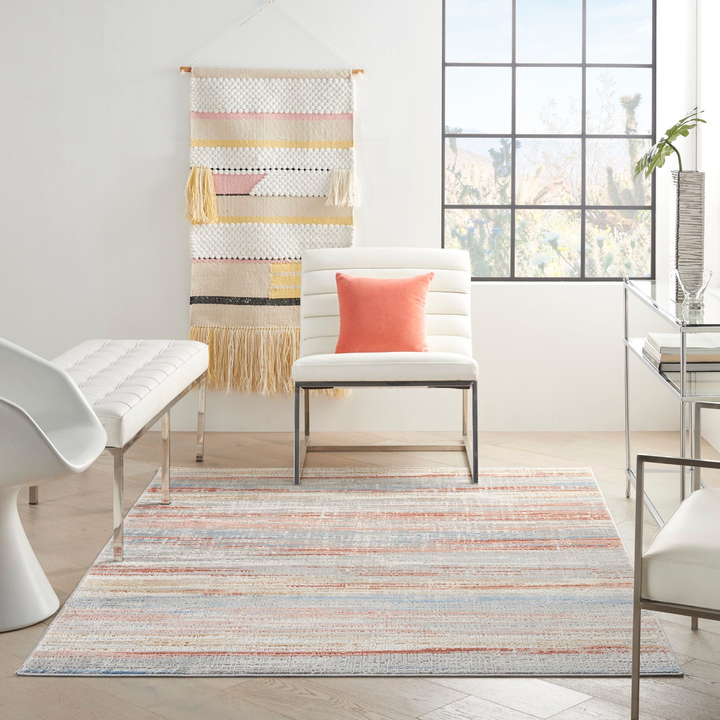 Bright modern living room with pastel striped rug, white leather chair, coral pillow, and a tassel-woven wall hanging.