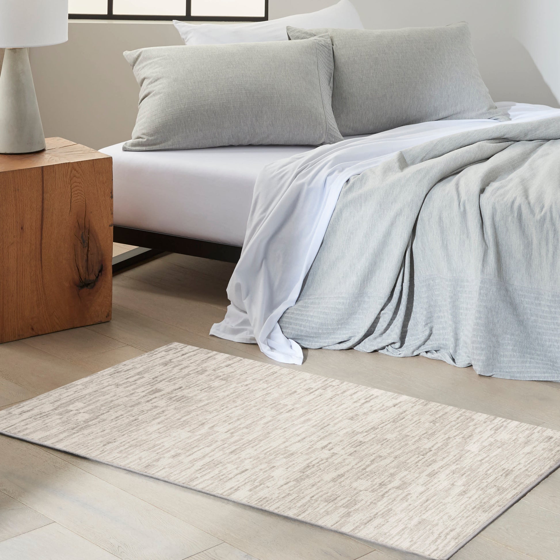 Rectangular beige textured rug with subtle pattern; light gray bedding and white sheets in a minimalist bedroom.